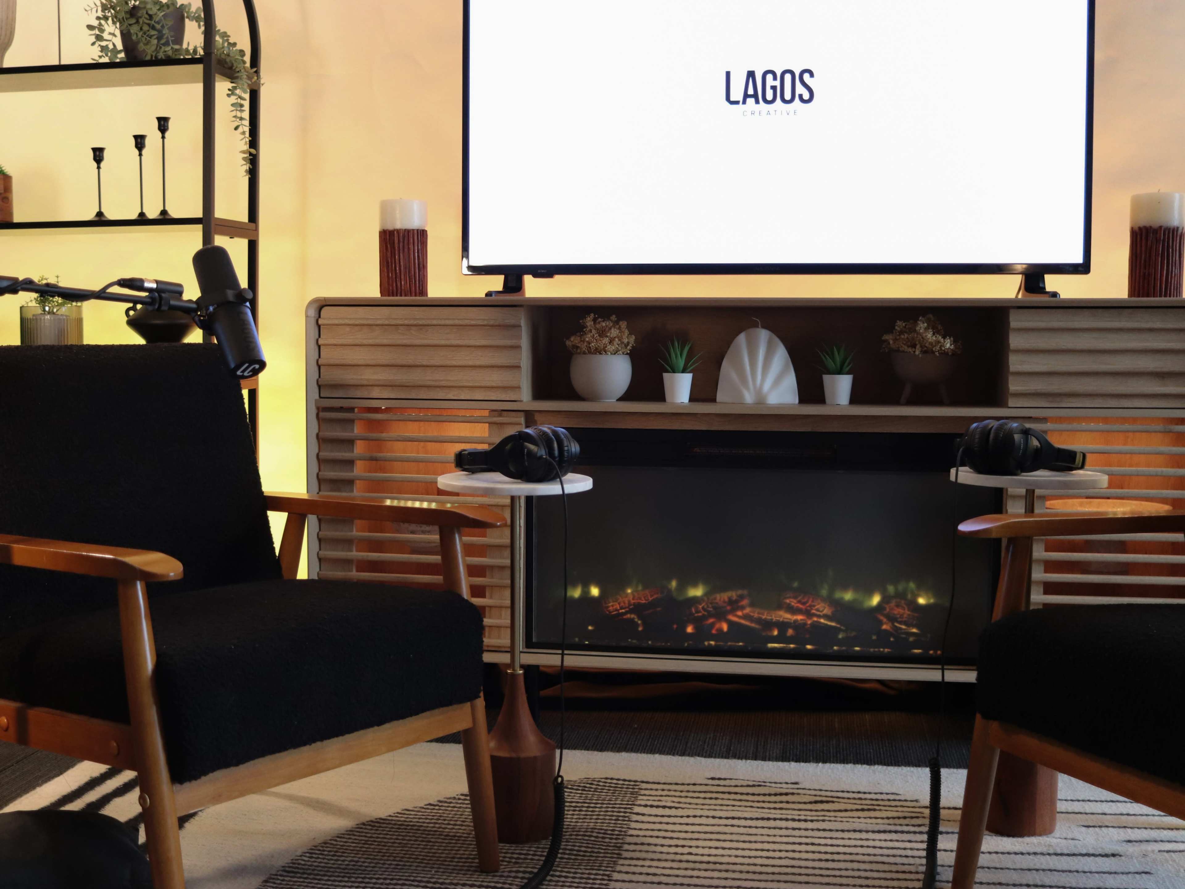 The image shows a cozy room setup featuring two black chairs facing a television displaying the text "LAGOS" on a wooden media console, with decorative plants and a fireplace below.