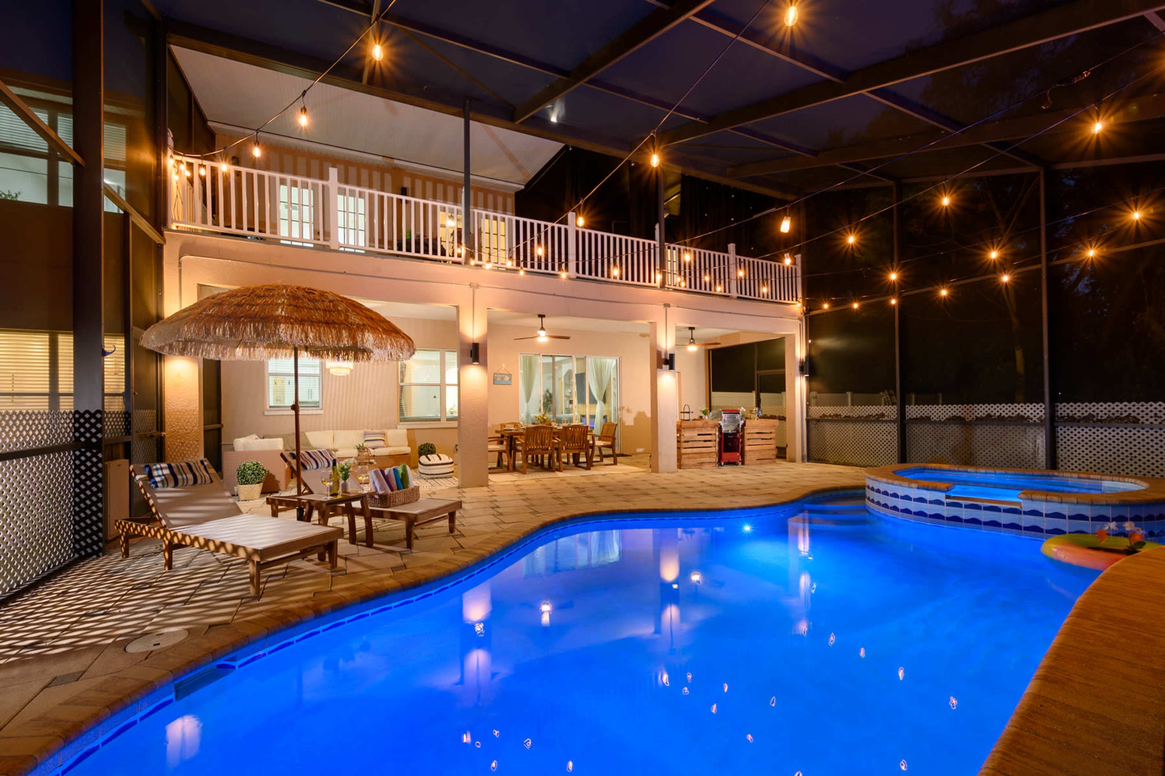 A well-lit outdoor pool area featuring a covered patio, dining space, and a hot tub surrounded by lounge chairs and decorative lights.