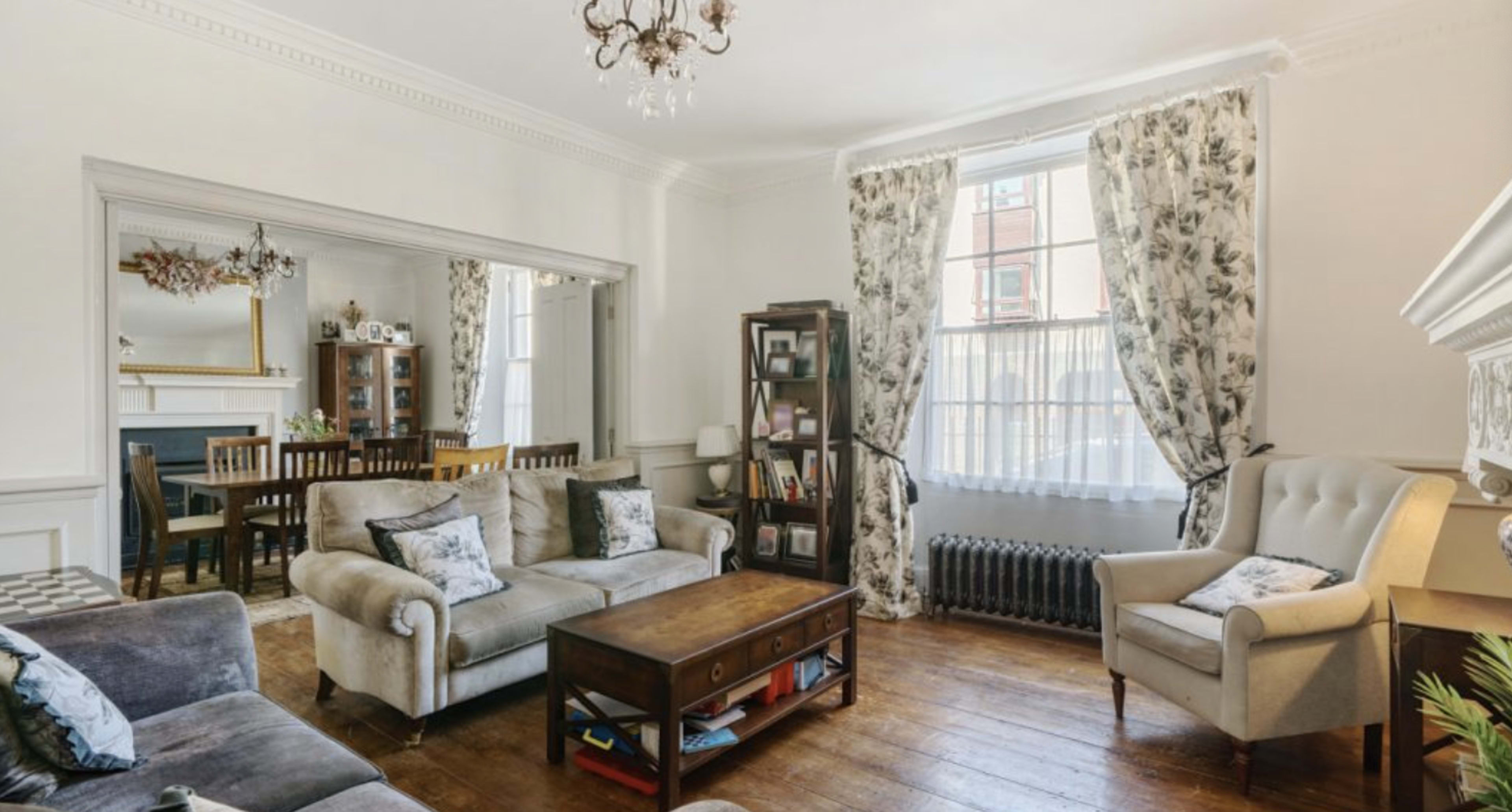 A spacious living room features a mix of light and dark furniture, a wooden coffee table, and large windows with floral curtains.