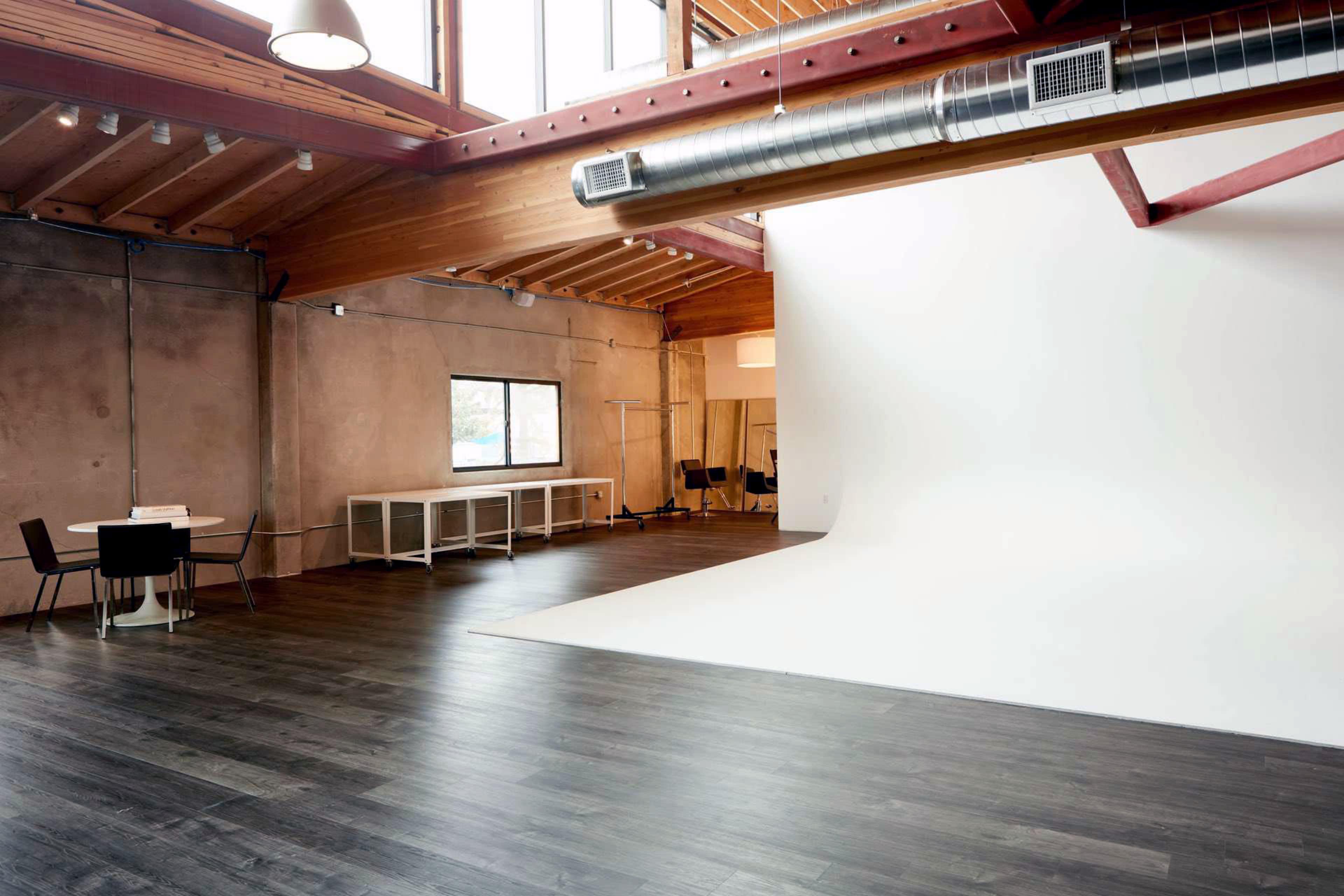 A spacious studio with wooden beams, a large white backdrop, and a seating area with a table and chairs.
