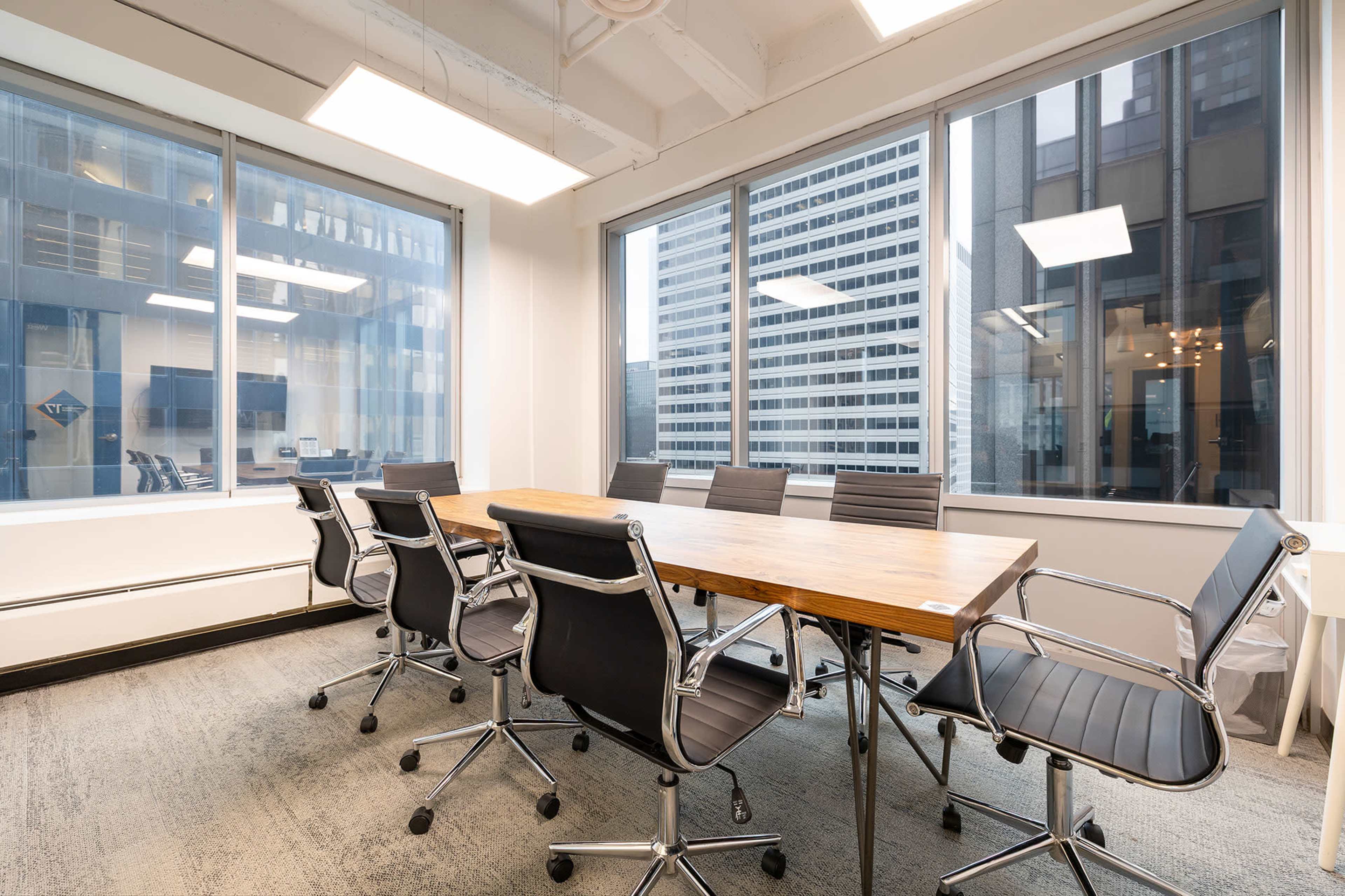 A conference room features a long wooden table surrounded by black rolling chairs, with large windows overlooking a cityscape.