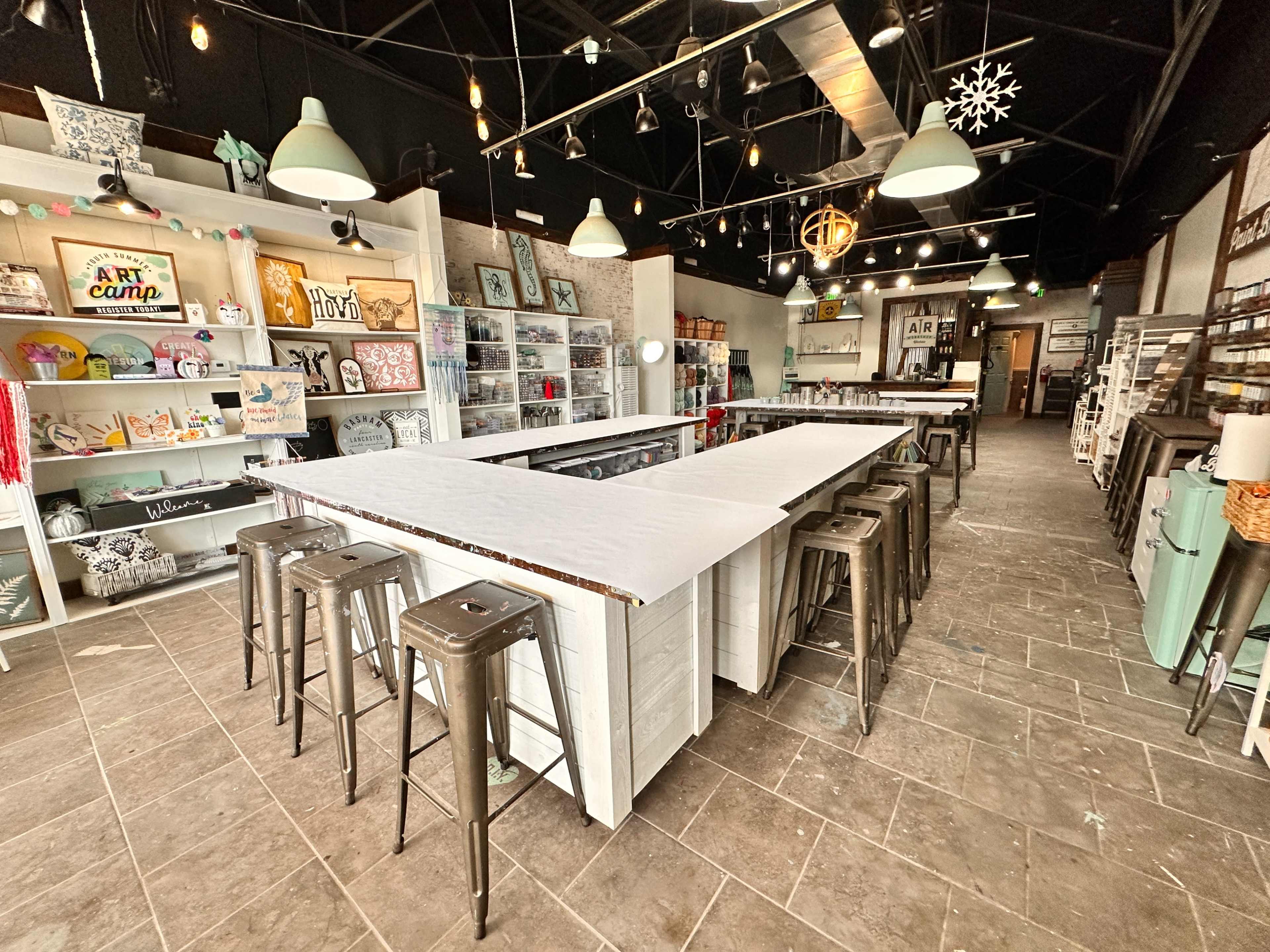An art studio features long white tables surrounded by metal stools, with shelves displaying various craft supplies and artworks.