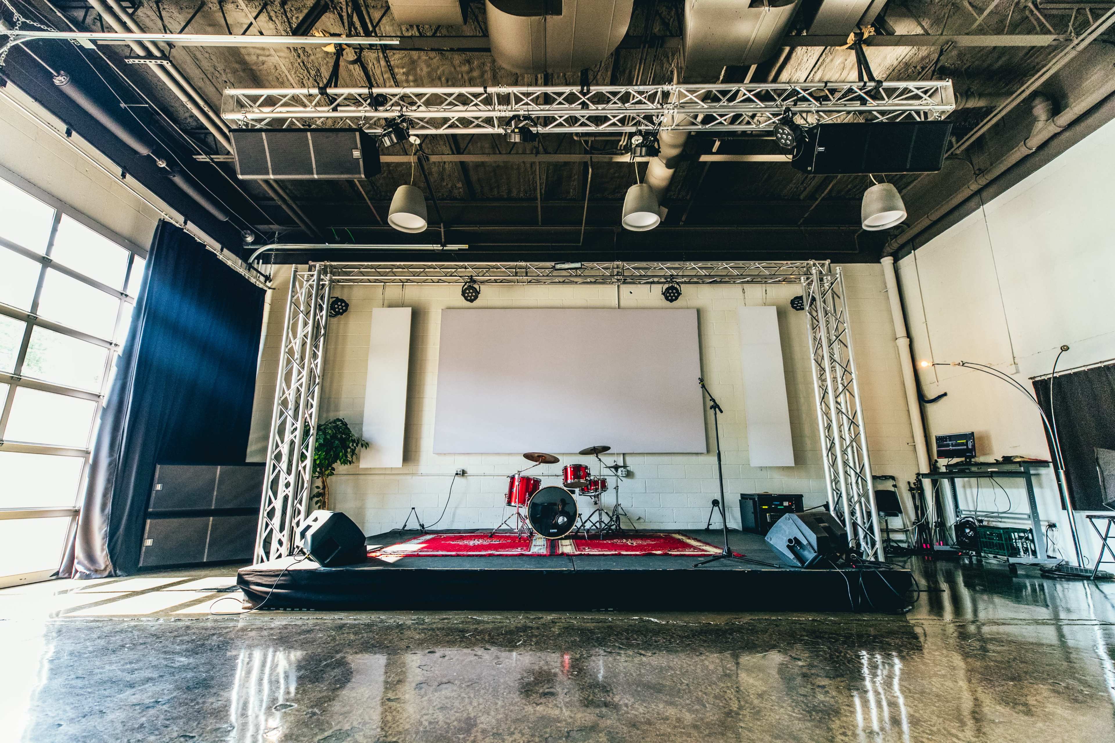 The image shows a stage set up with a drum kit, sound equipment, and a large screen in a spacious, well-lit venue.