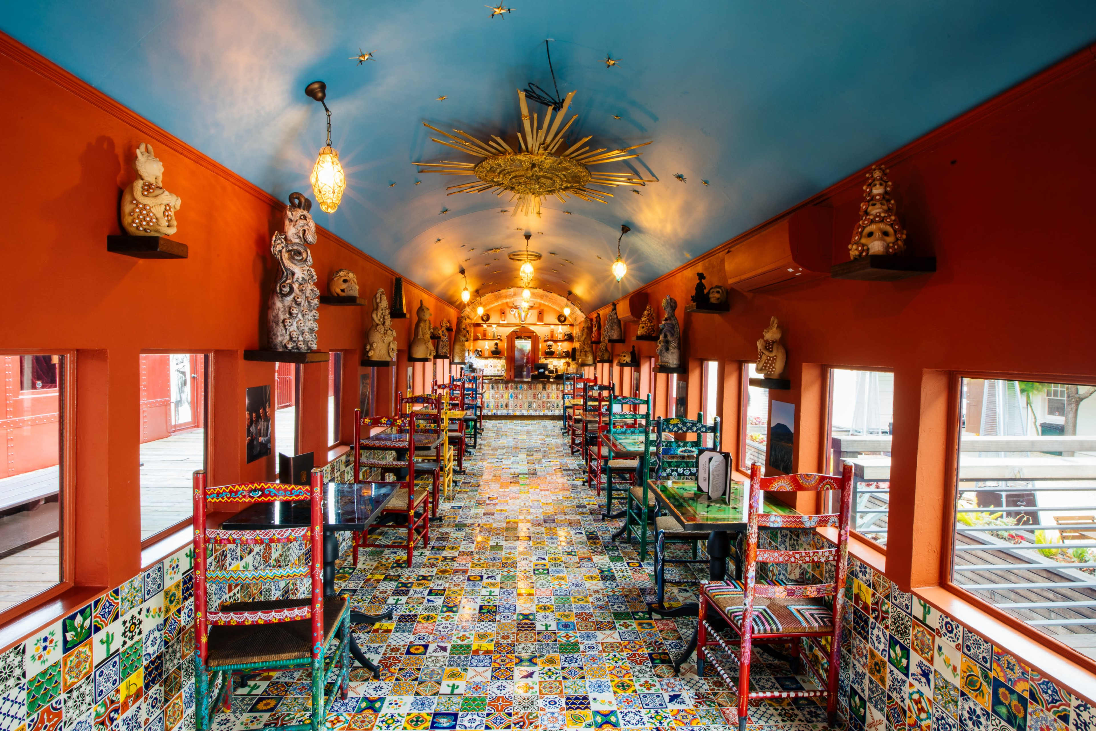The image shows a brightly colored dining area with a patterned tile floor, adorned with various decorative figurines on the walls and colorful chairs around the tables.