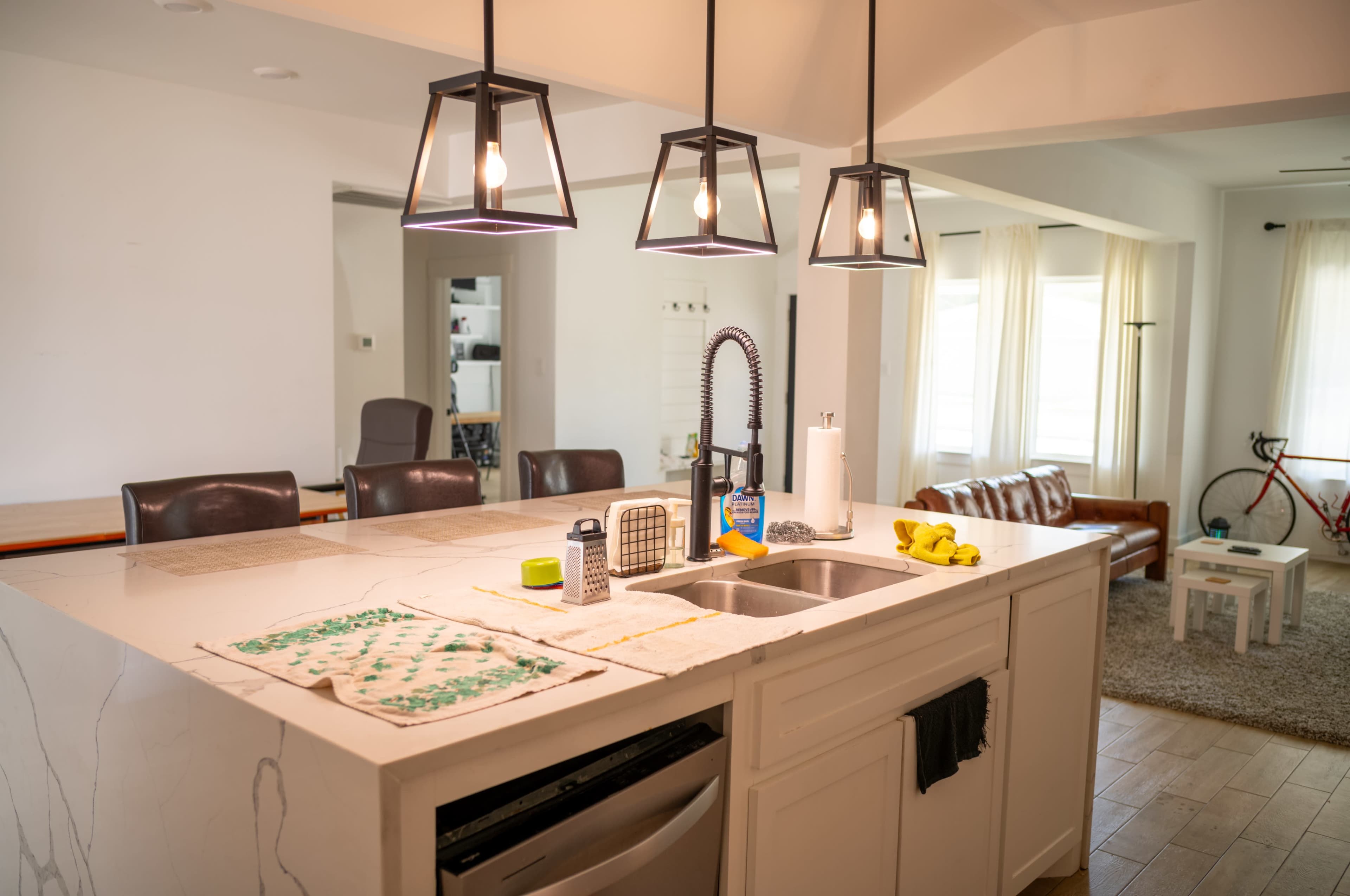 A modern kitchen features a central island with a sink, surrounded by pendant lights, and a living area visible in the background with a bicycle and seating.