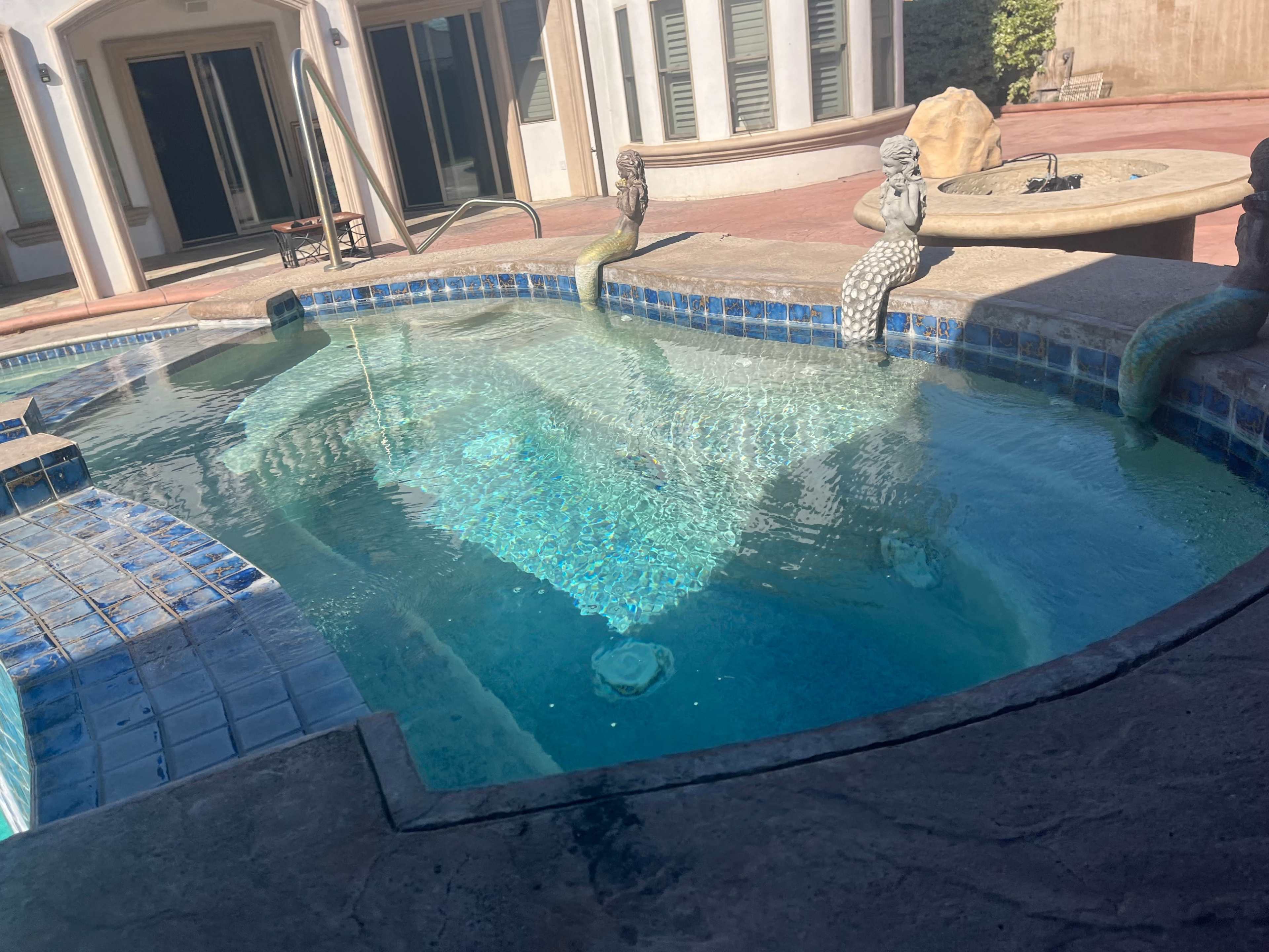 A decorative swimming pool features a stone mermaid sculpture and a fountain, surrounded by tiled edges and a patio area.