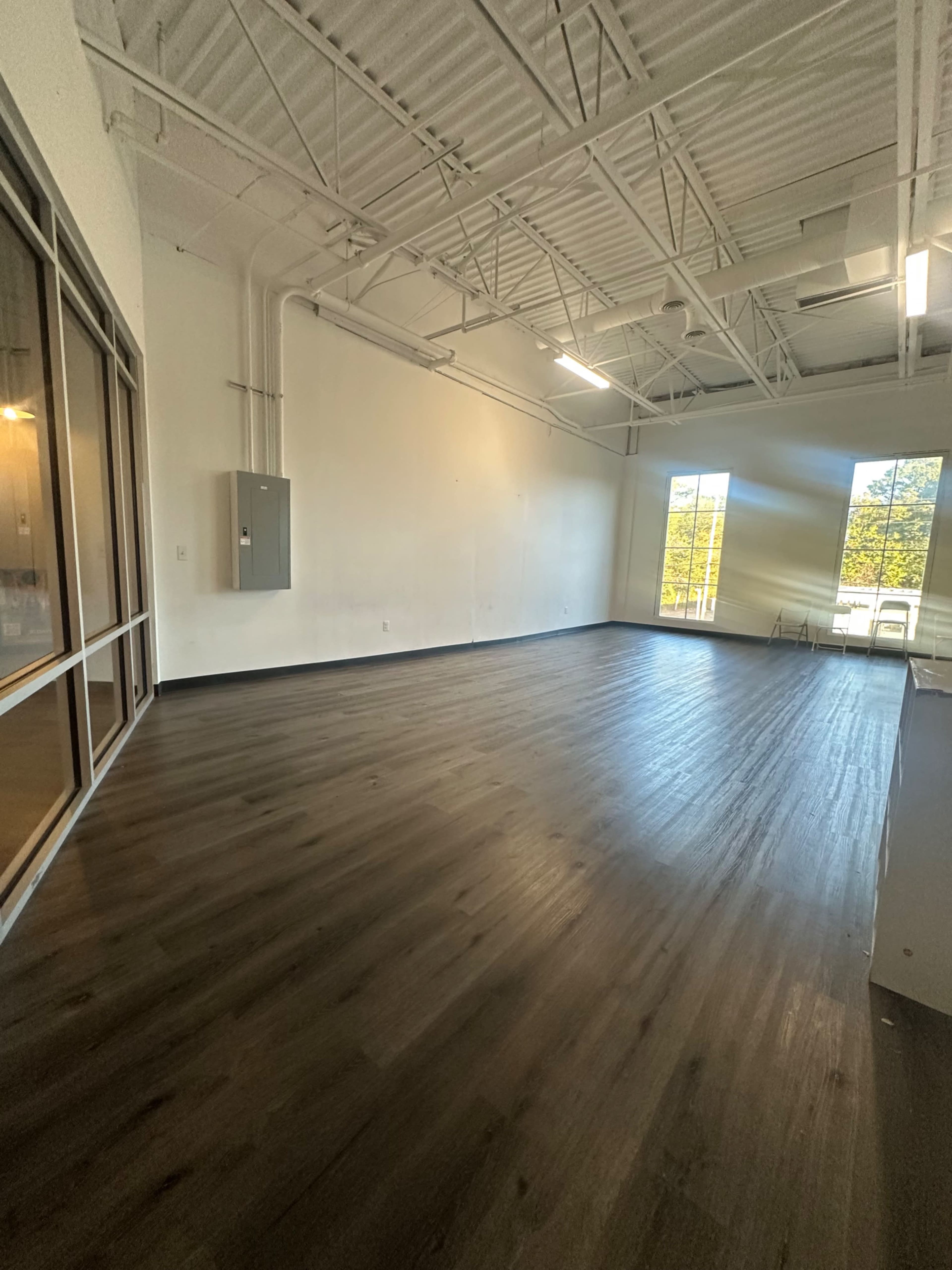 The image shows an empty room with a wooden floor, large windows on one wall, and white walls.