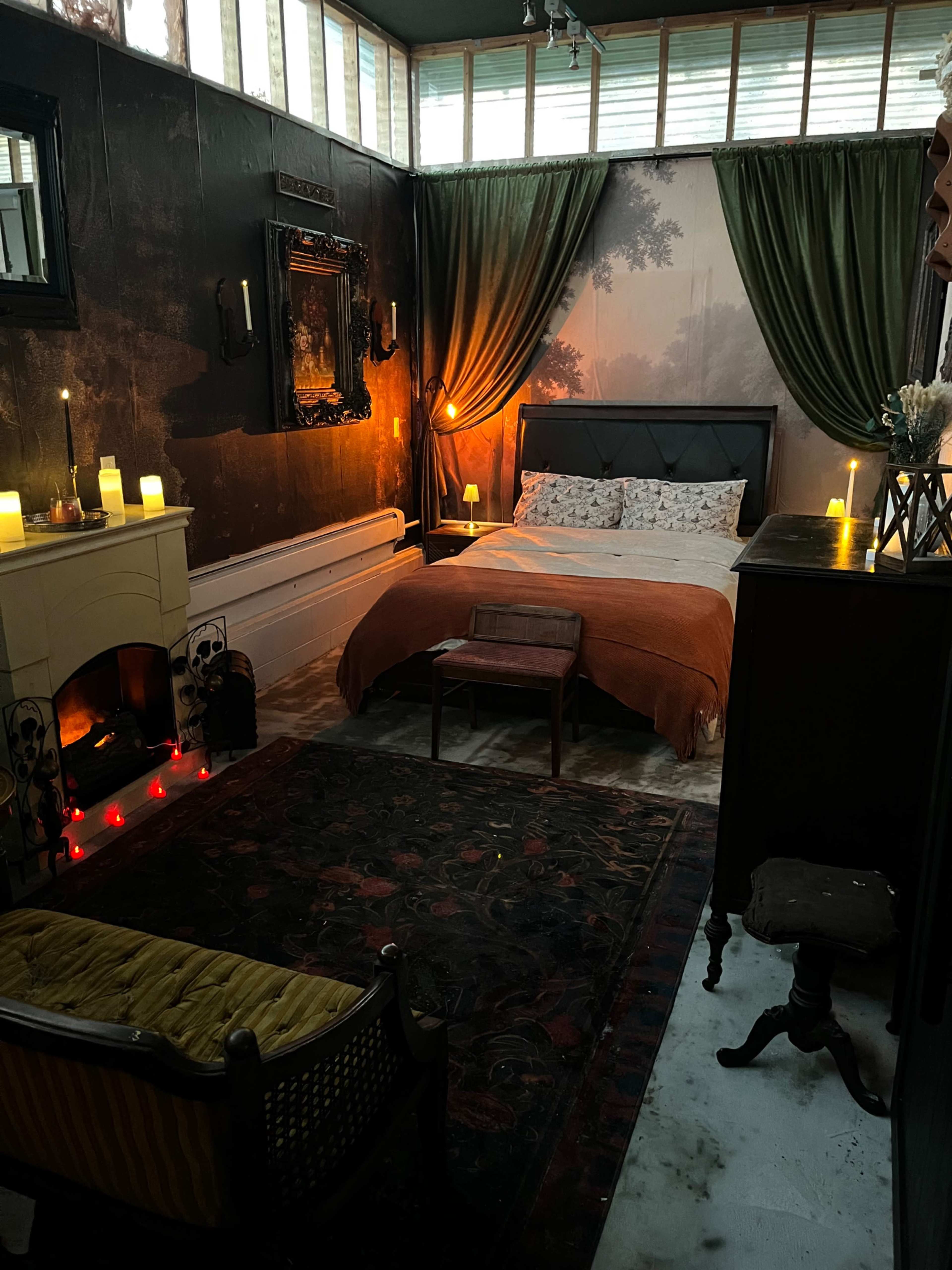 The image shows a dimly lit bedroom featuring a bed with an orange throw, decorative curtains, a fireplace with candles, and vintage furniture.