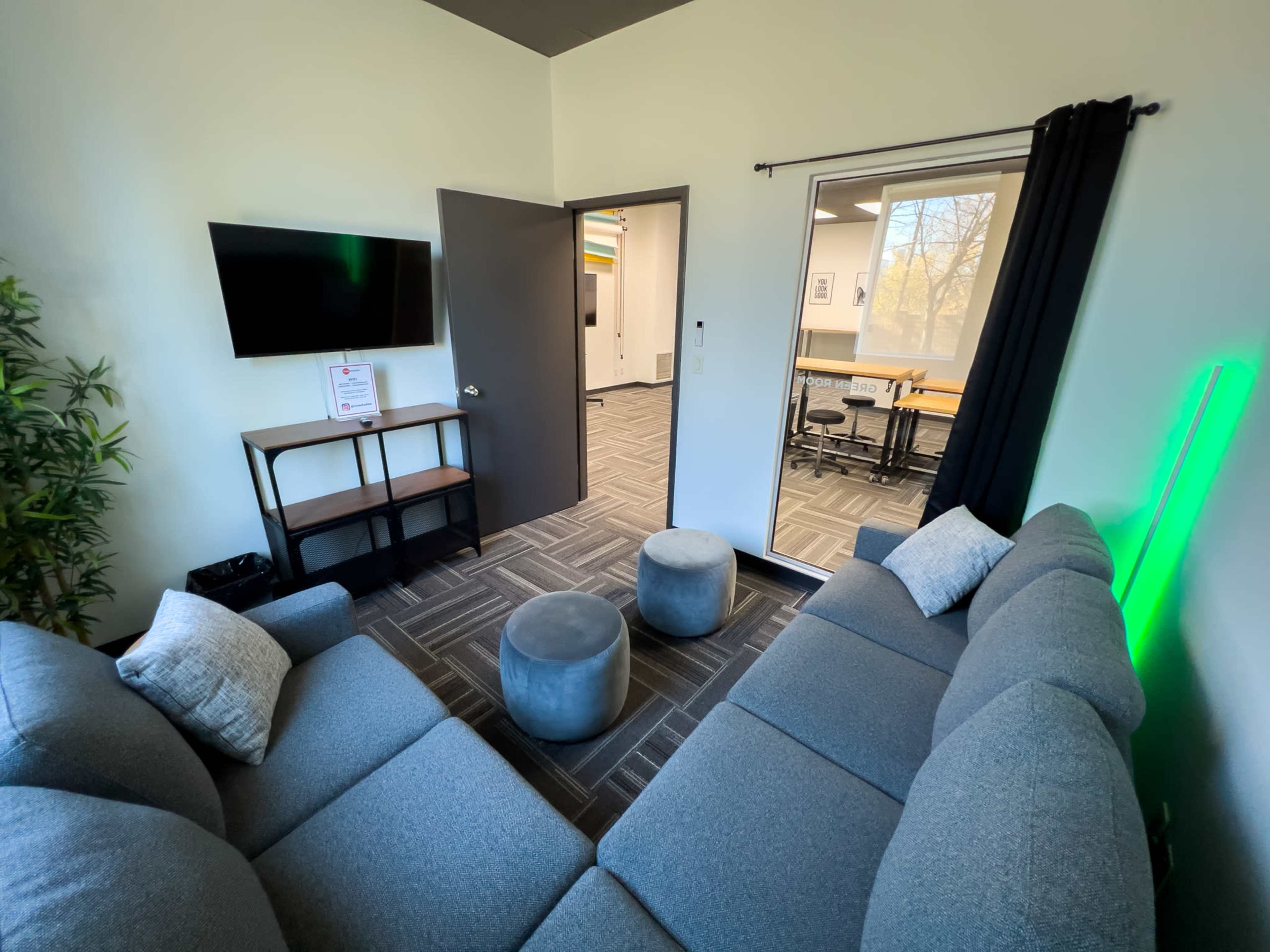 A modern small lounge area featuring a gray sectional sofa, a television on a stand, and a round ottoman, with an open door leading to a conference room.