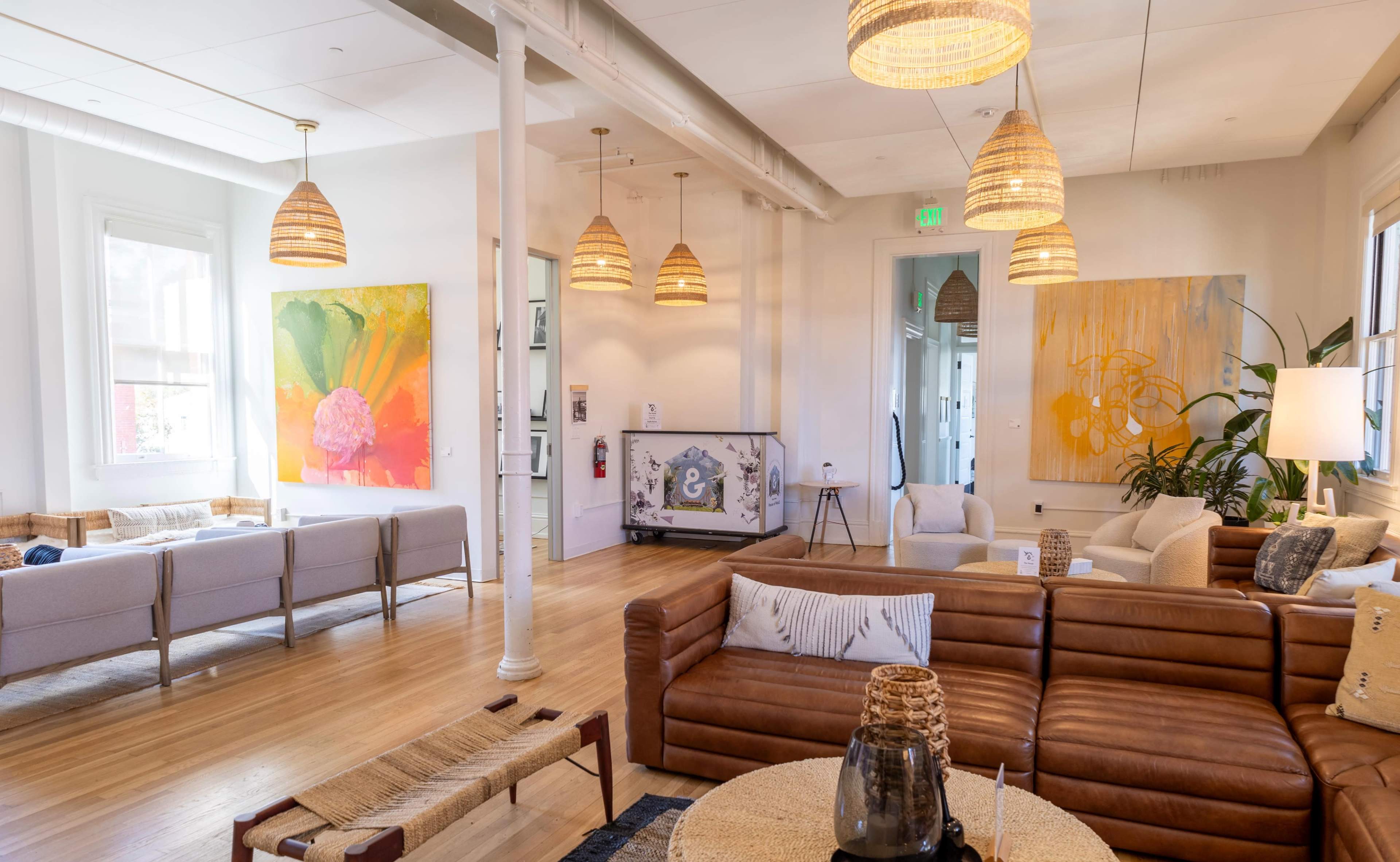 The image shows a bright, modern living space featuring a leather sofa, a woven coffee table, and large, colorful wall art.