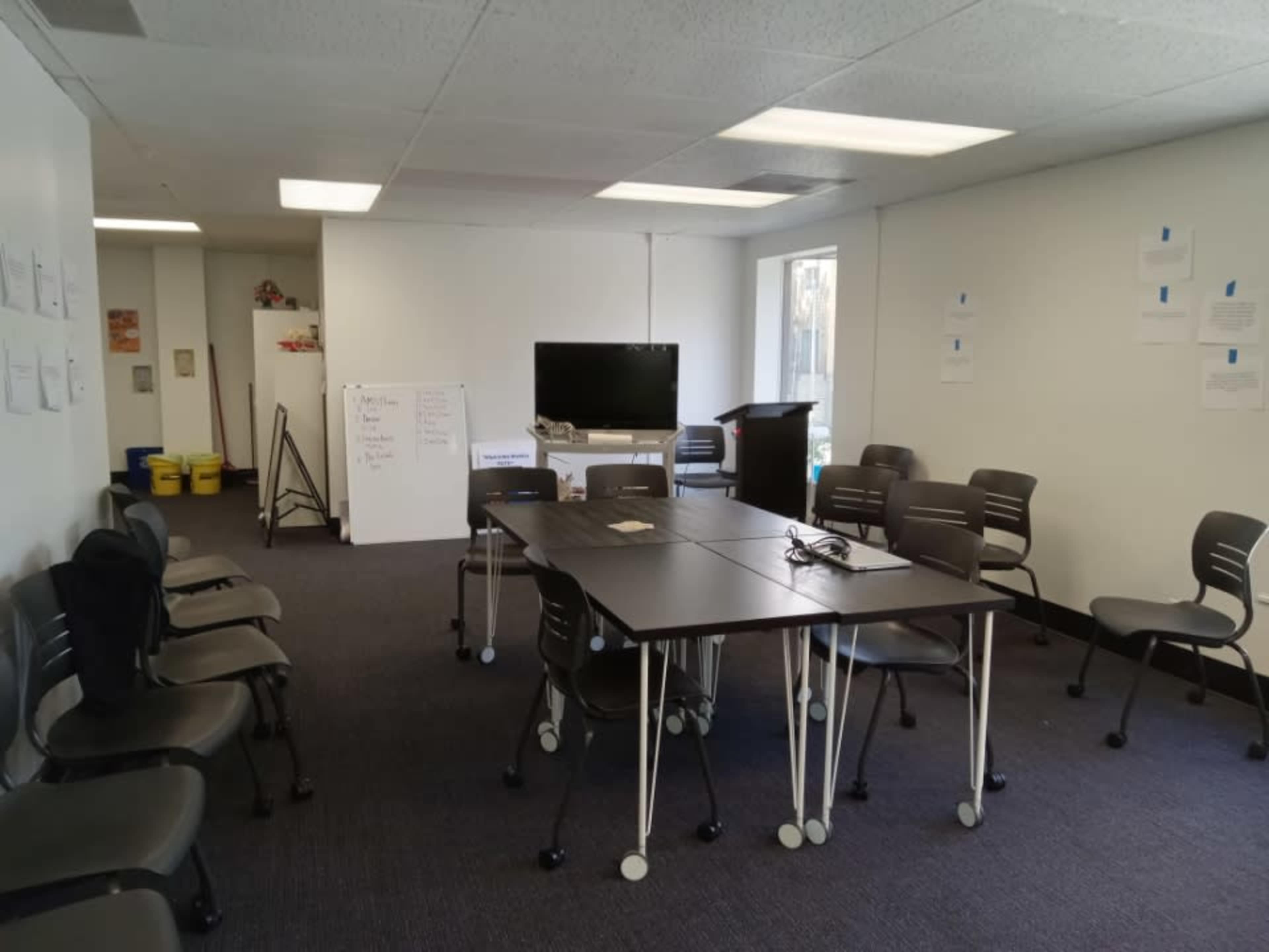 A conference room is set up with a rectangular table surrounded by chairs, a television on a stand, and whiteboards against the walls.
