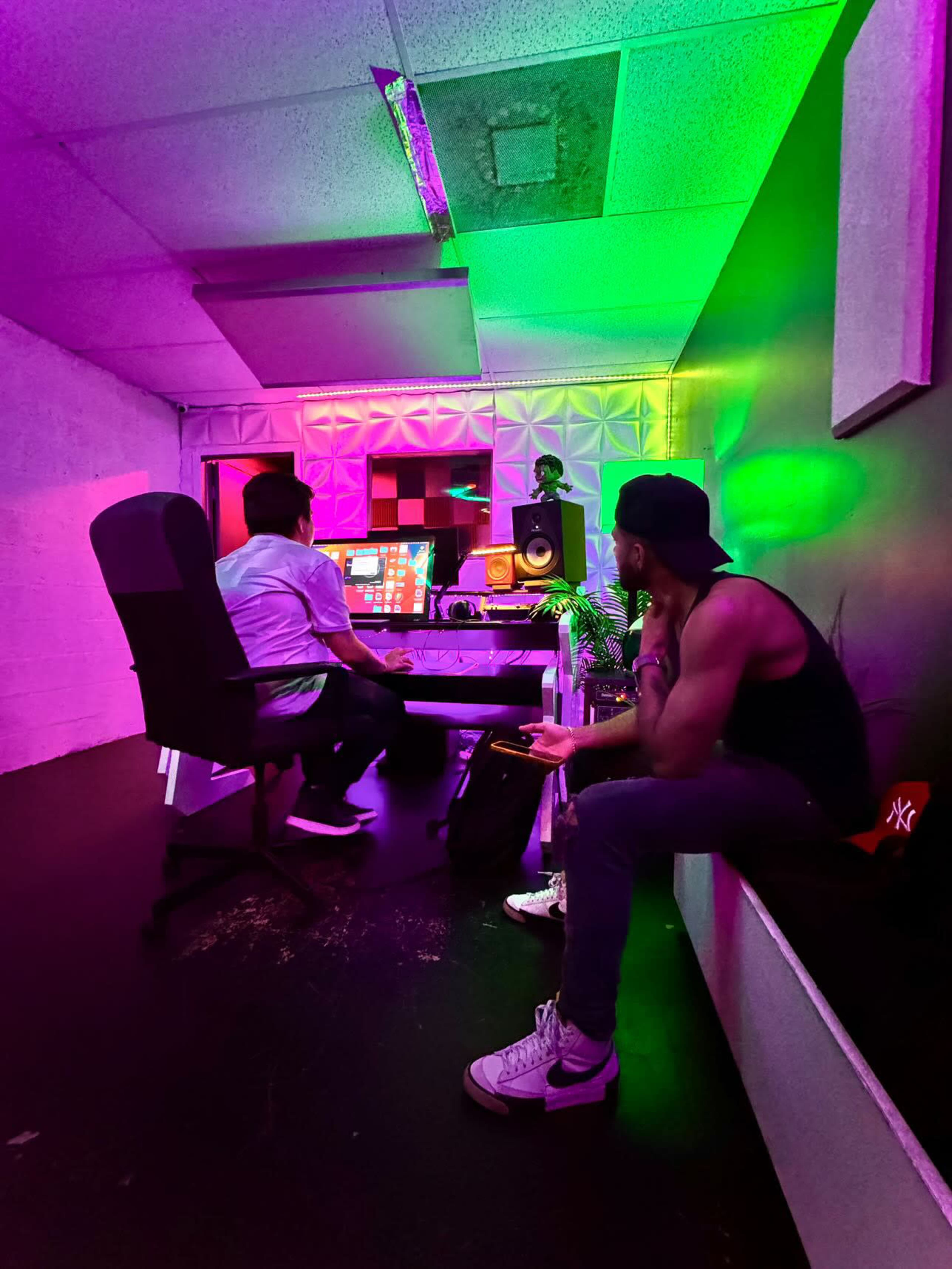 Two individuals are seated in a music studio, with colorful neon lighting illuminating the room as one works on a music production setup while the other observes.