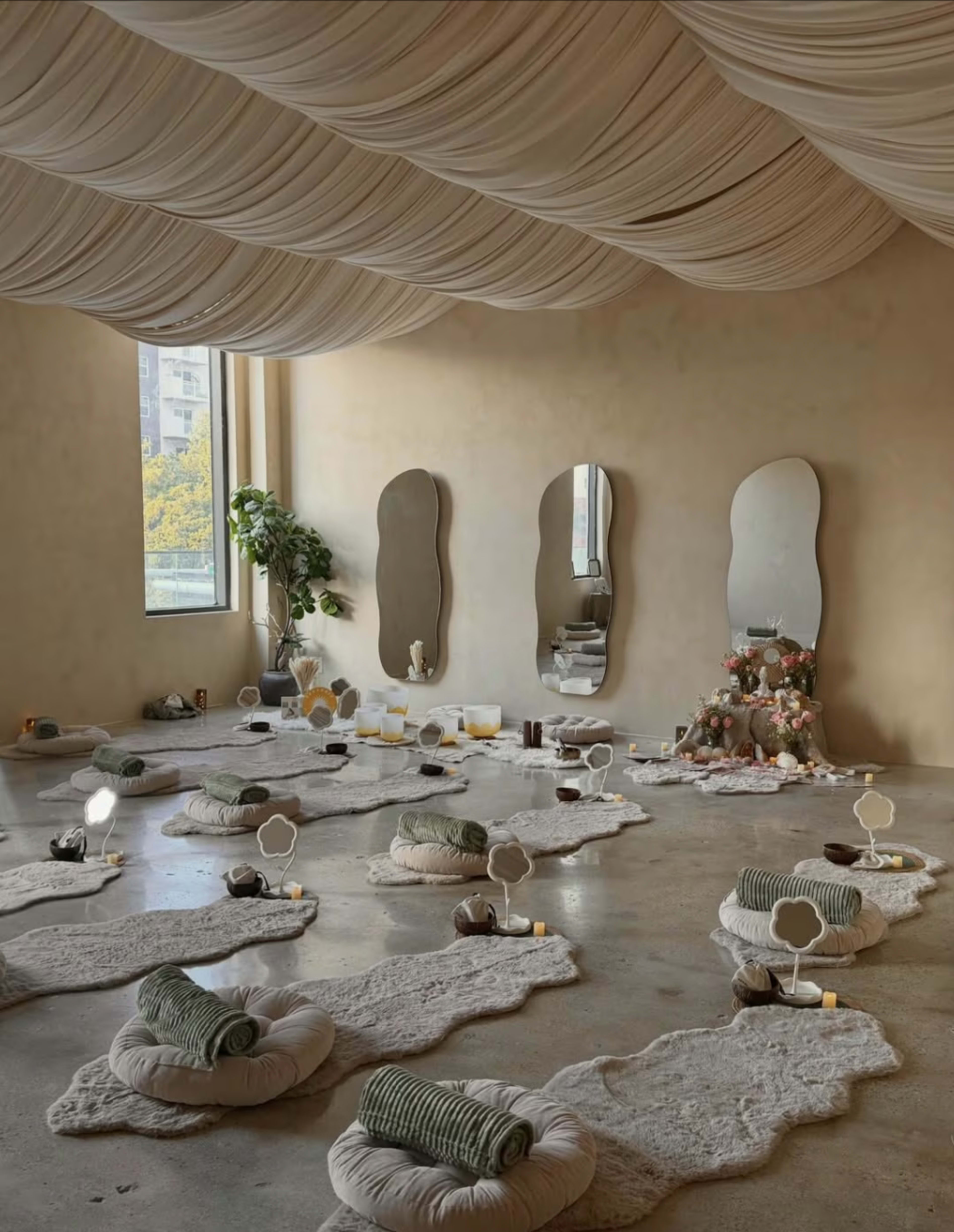 The image shows a serene indoor space designed for relaxation, featuring individual meditation areas with cushions, mirrors, and soft lighting against a neutral-colored wall.