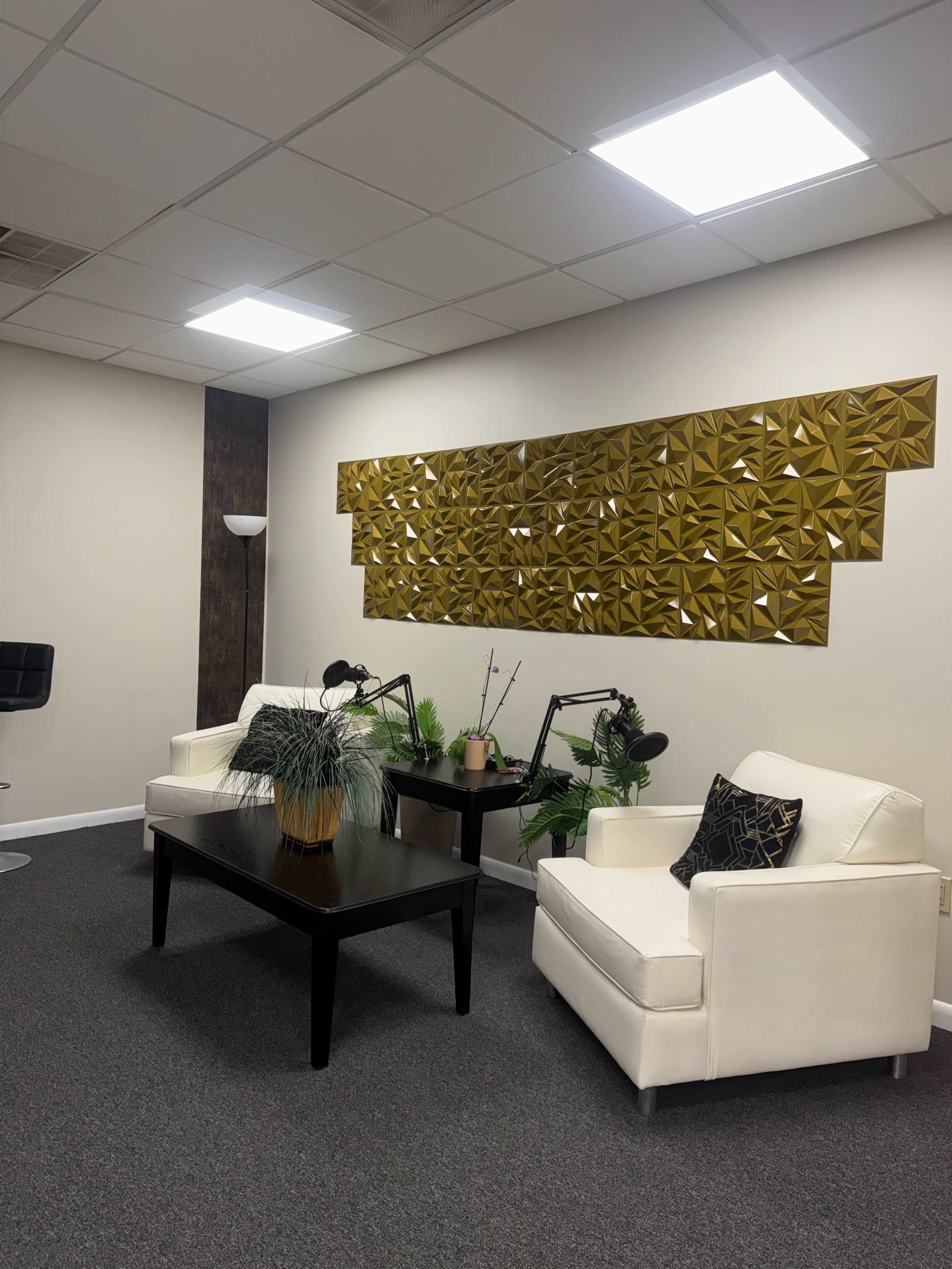 The image shows a modern office space featuring two white armchairs, a black table, and a decorative gold wall panel.
