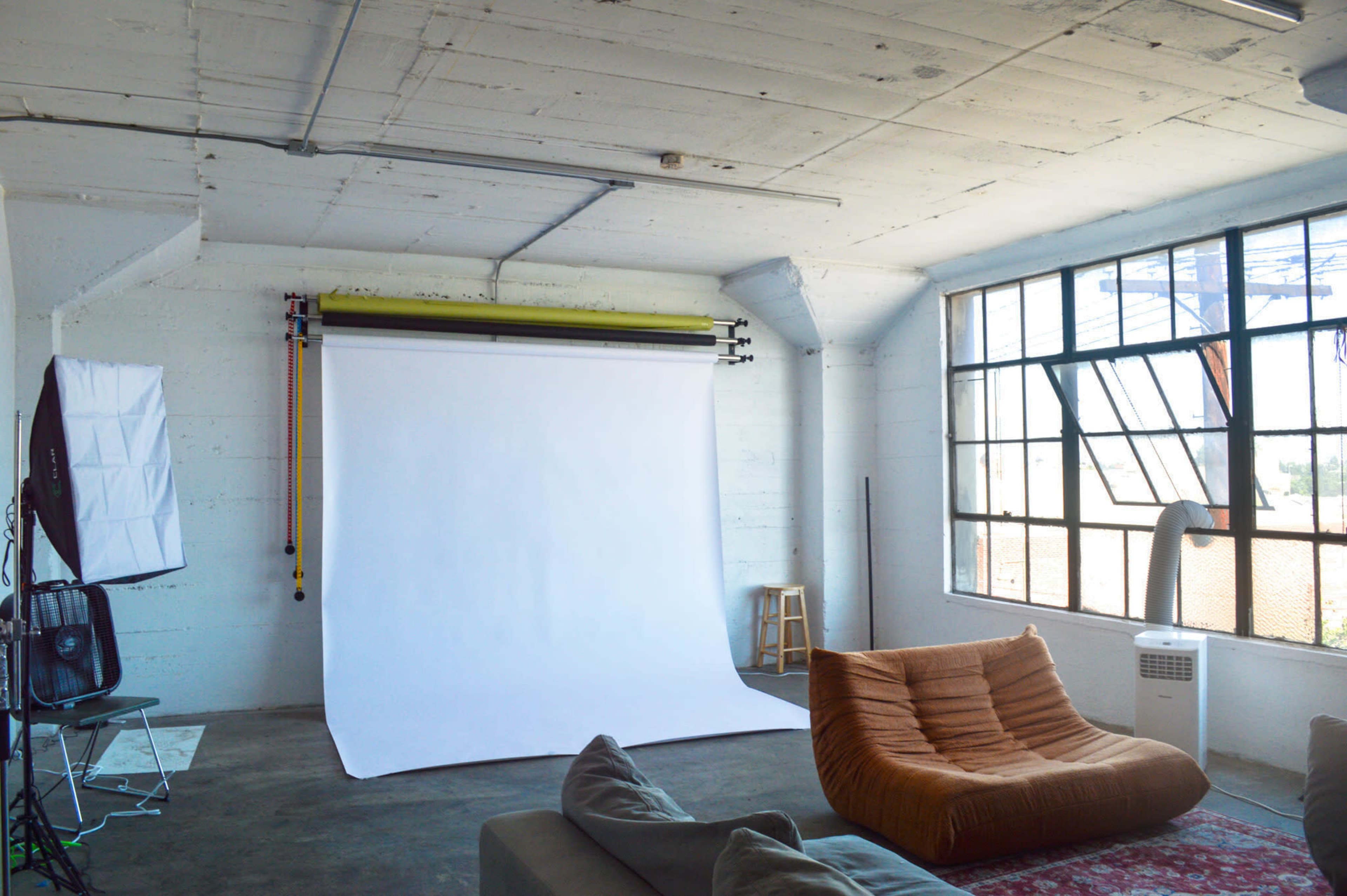 A photography studio features a large white backdrop, a comfortable couch, and studio lighting equipment arranged in a well-lit space with industrial windows.