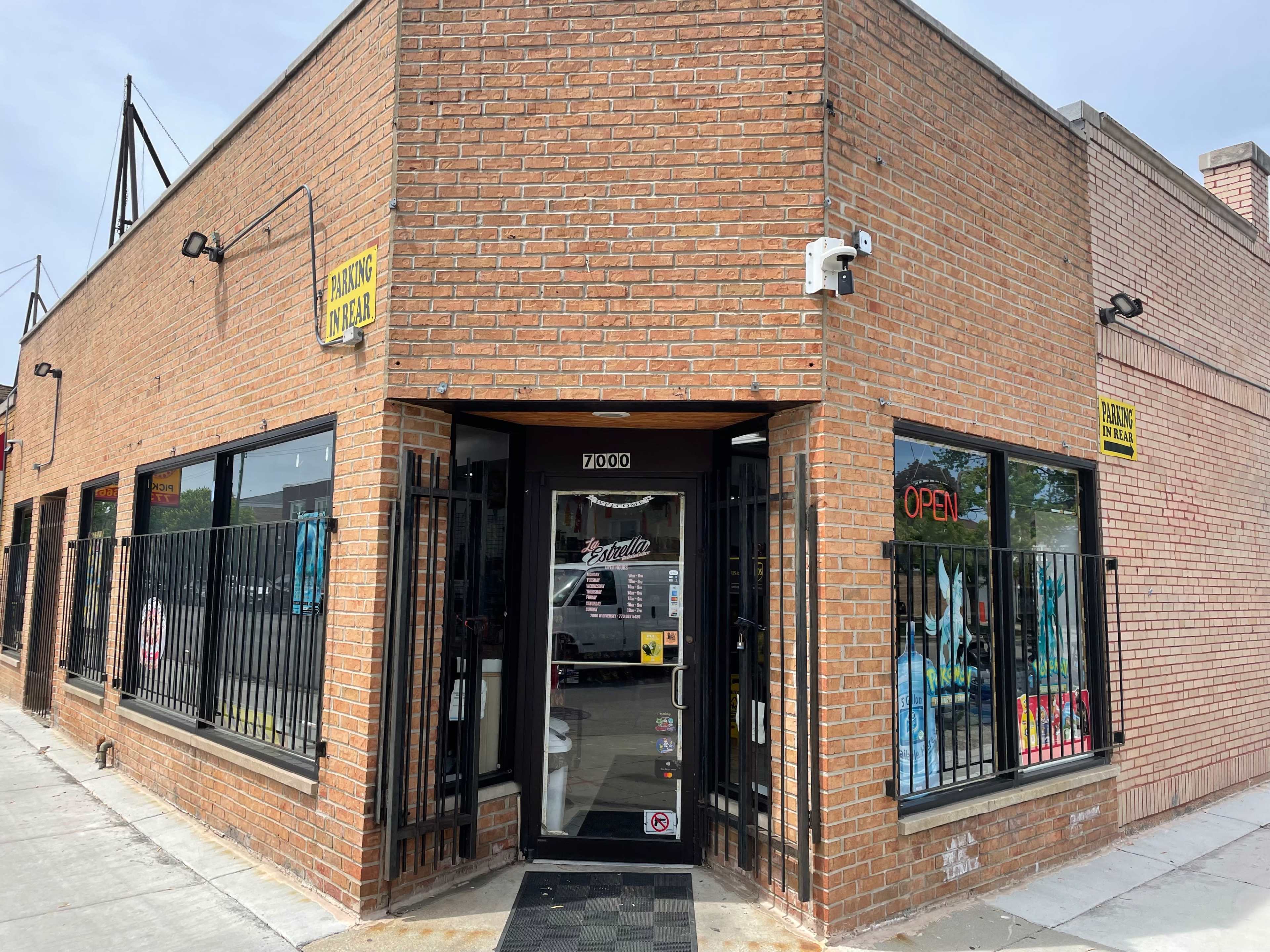 A brick storefront with large windows displays an "OPEN" sign and is secured with a black metal gate.