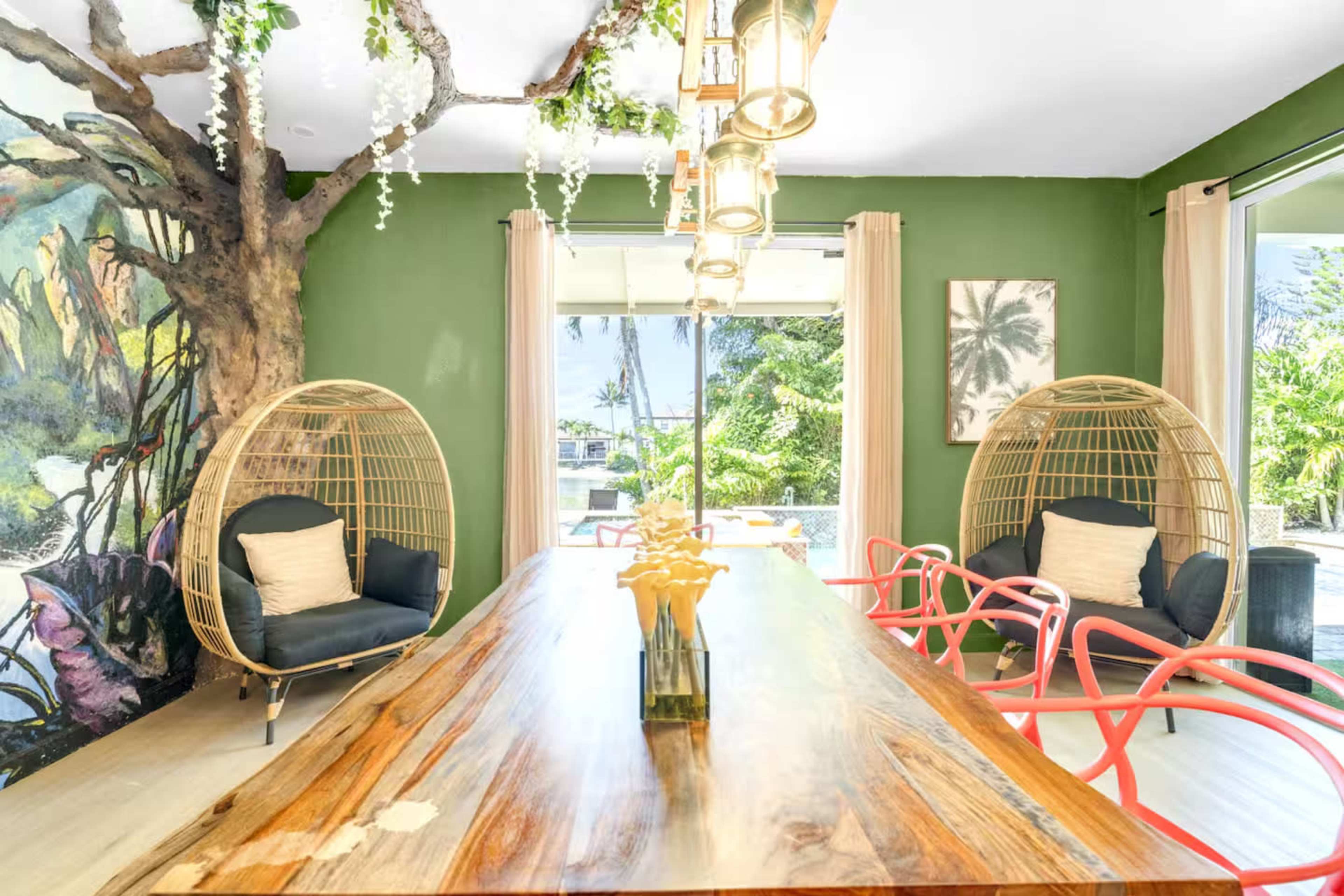 The image shows a dining area with a wooden table surrounded by two rattan chairs and colorful red chairs, against a green wall with a large mural and a view of outdoor greenery through glass doors.
