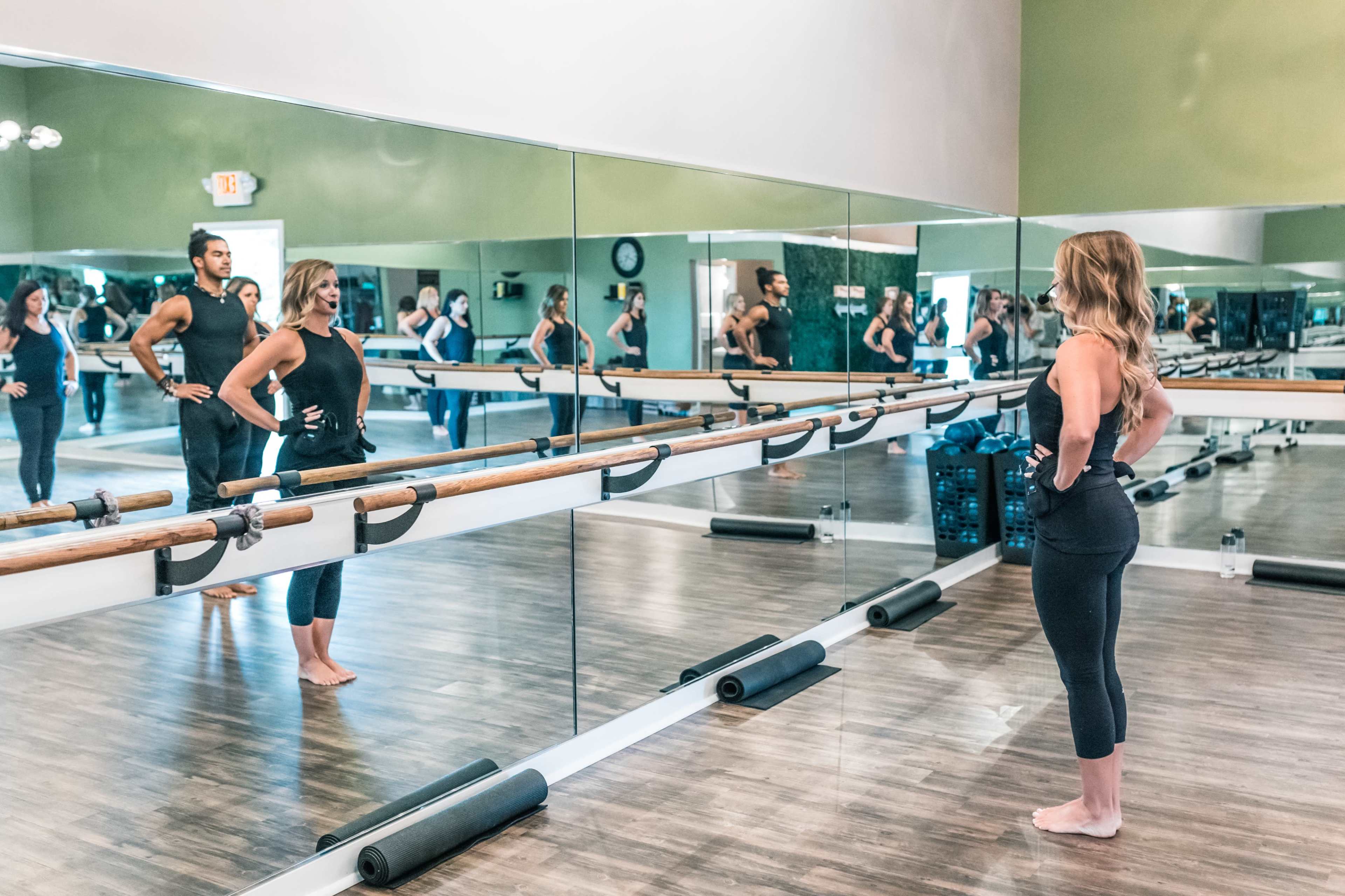 A group of people practices fitness at a studio with a mirrored wall and wooden barre.