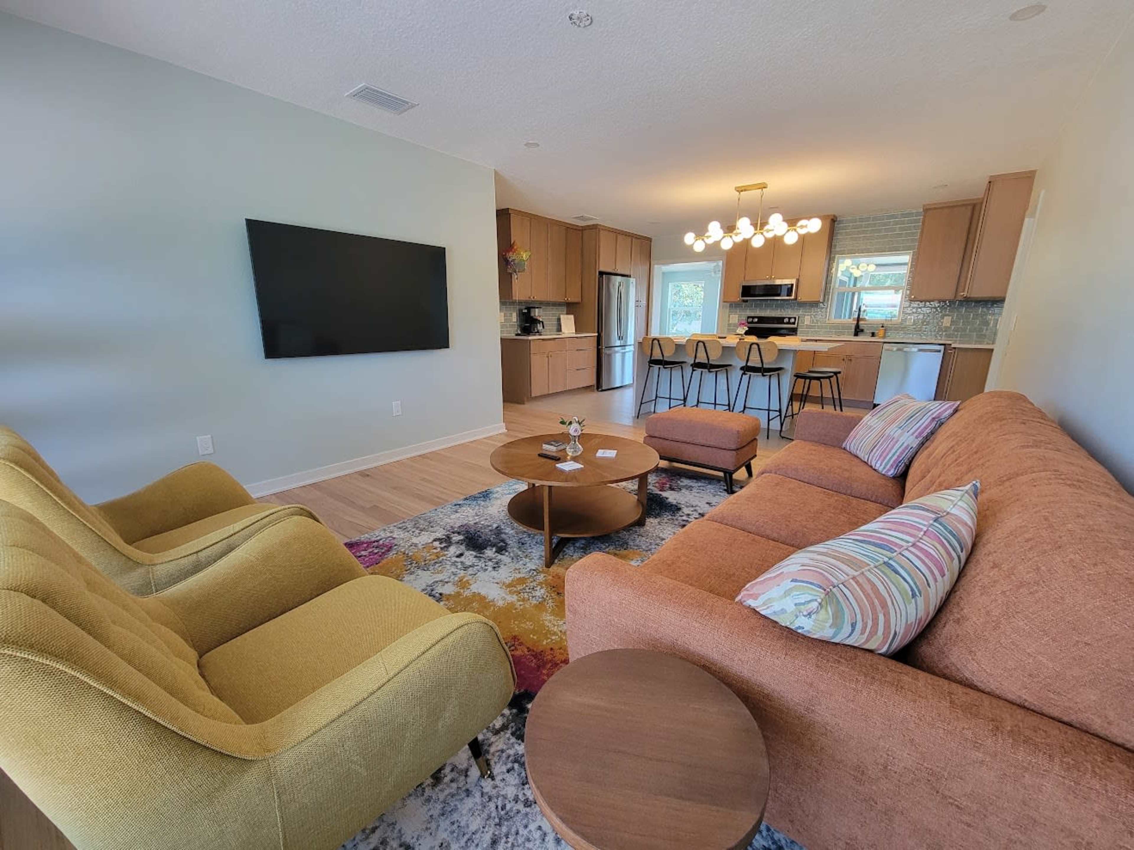 The image depicts a modern living room with a brown sectional sofa, two yellow accent chairs, a coffee table, and a wall-mounted television, leading to an open kitchen area with bar seating.