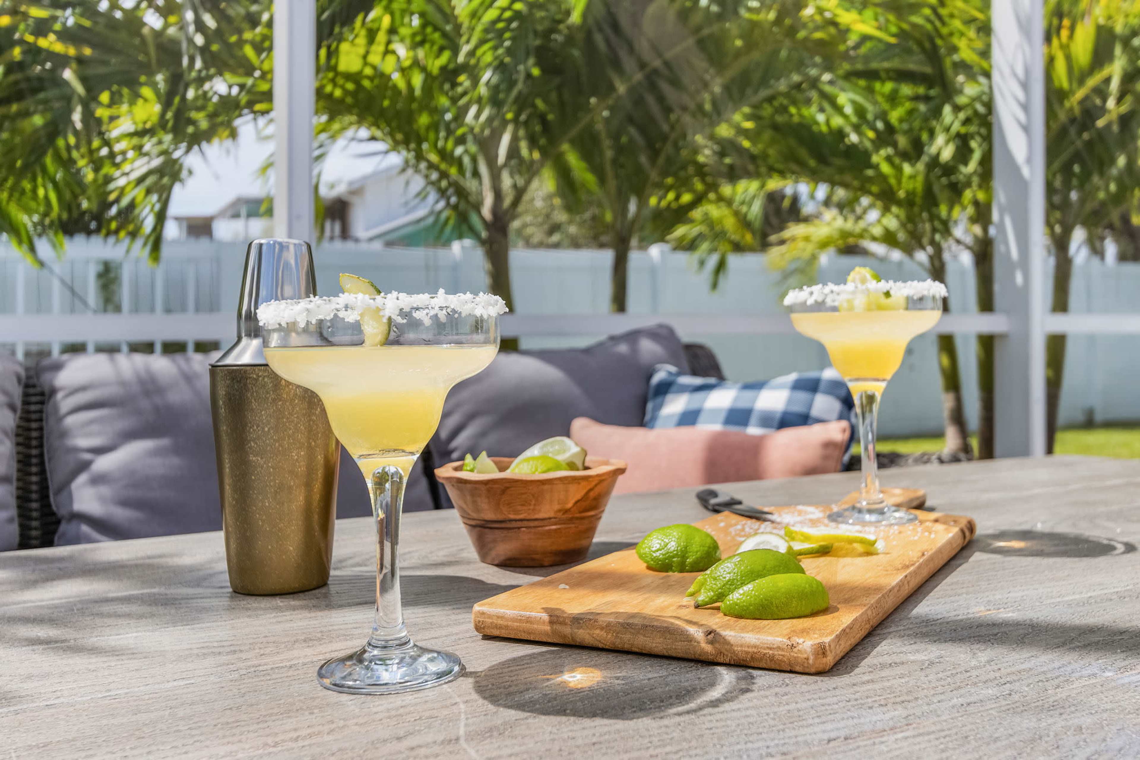 Two margarita cocktails with salt-rimmed glasses sit on a table alongside a wooden cutting board with limes and a cocktail shaker, set against a backdrop of palm trees.