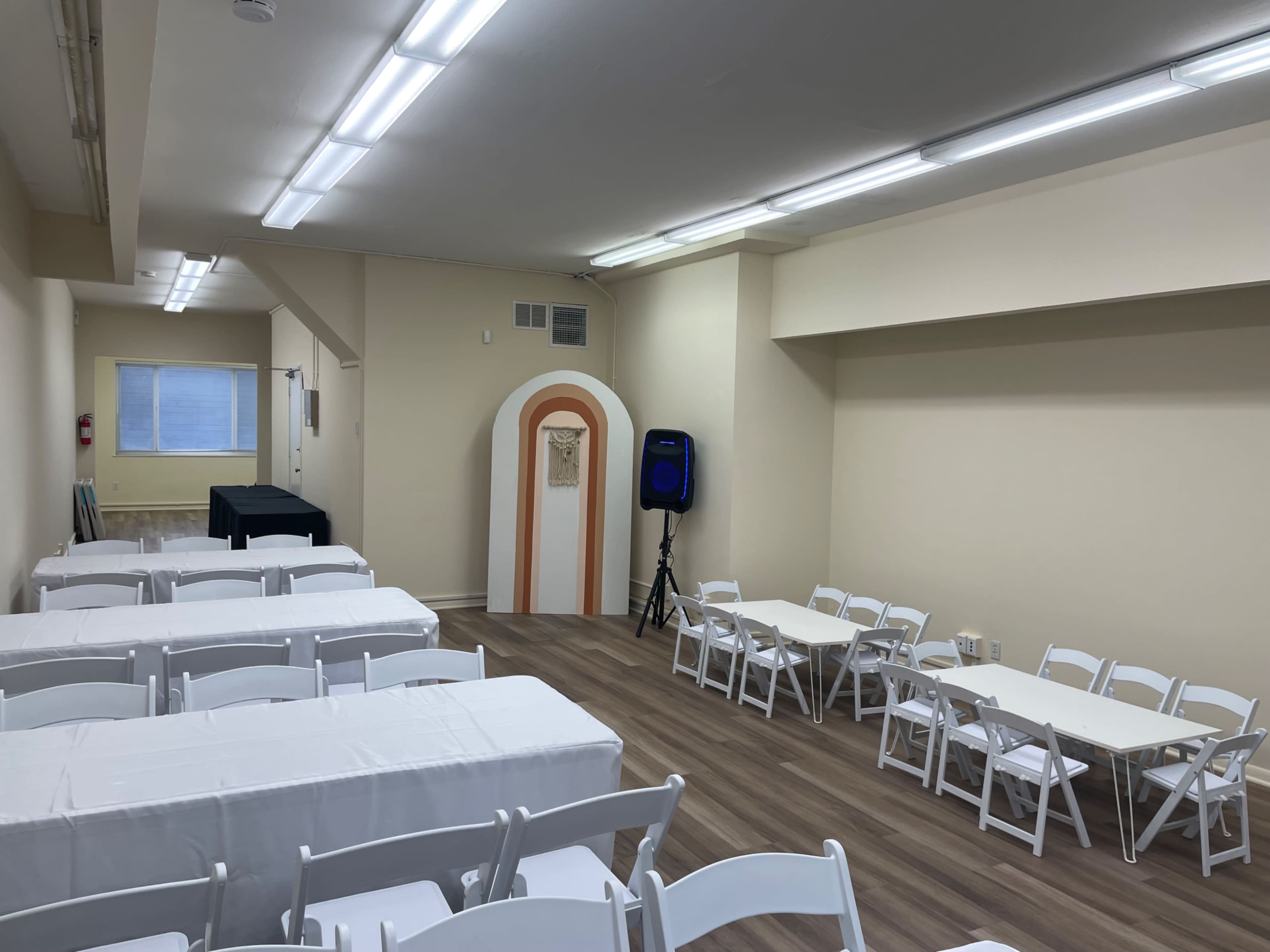 A spacious room set up with several white tables and chairs arranged for a gathering, with a doorway featuring an arch in the background.