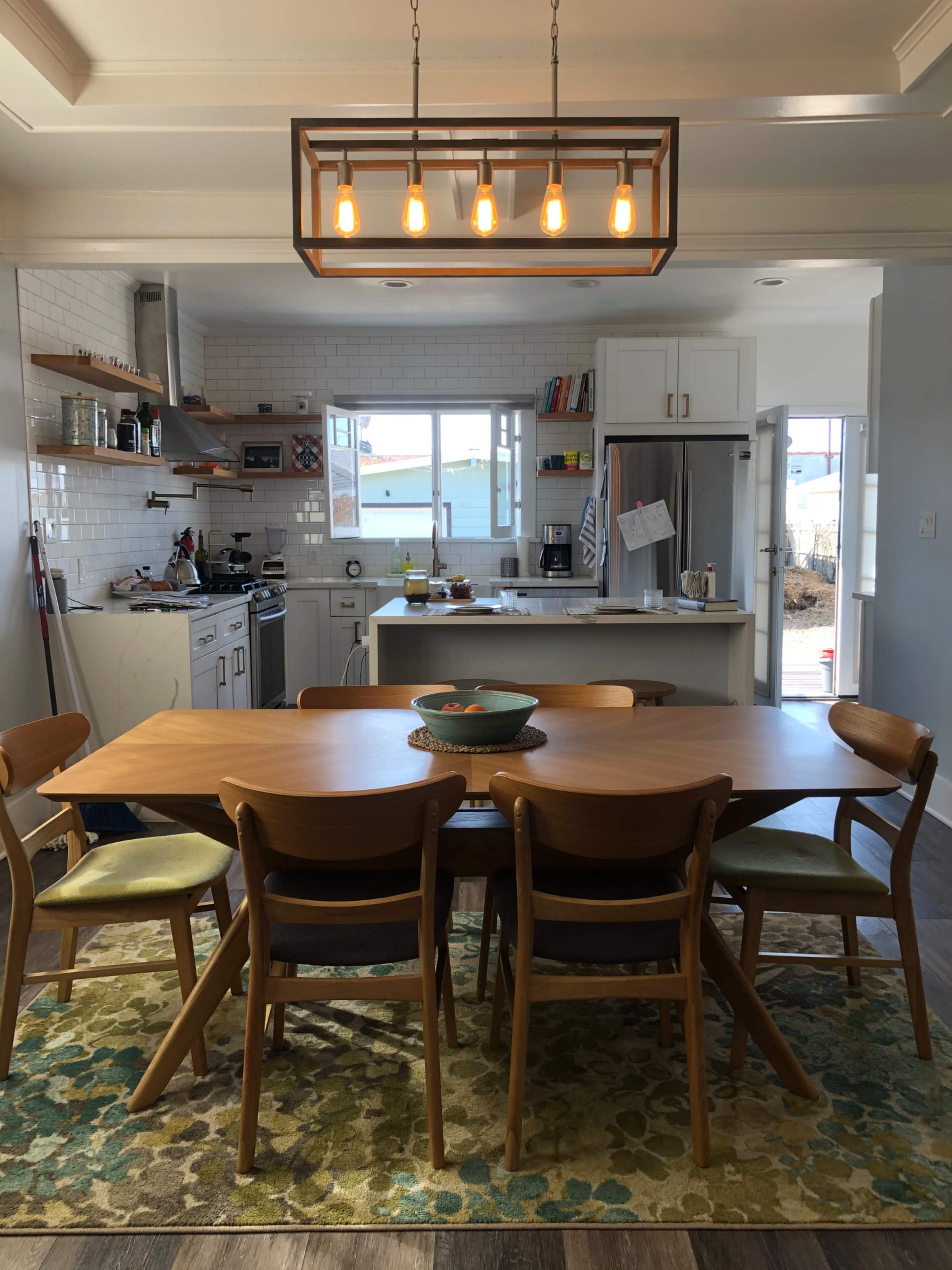 A wooden dining table surrounded by chairs is positioned in a kitchen with modern appliances and a light fixture overhead.