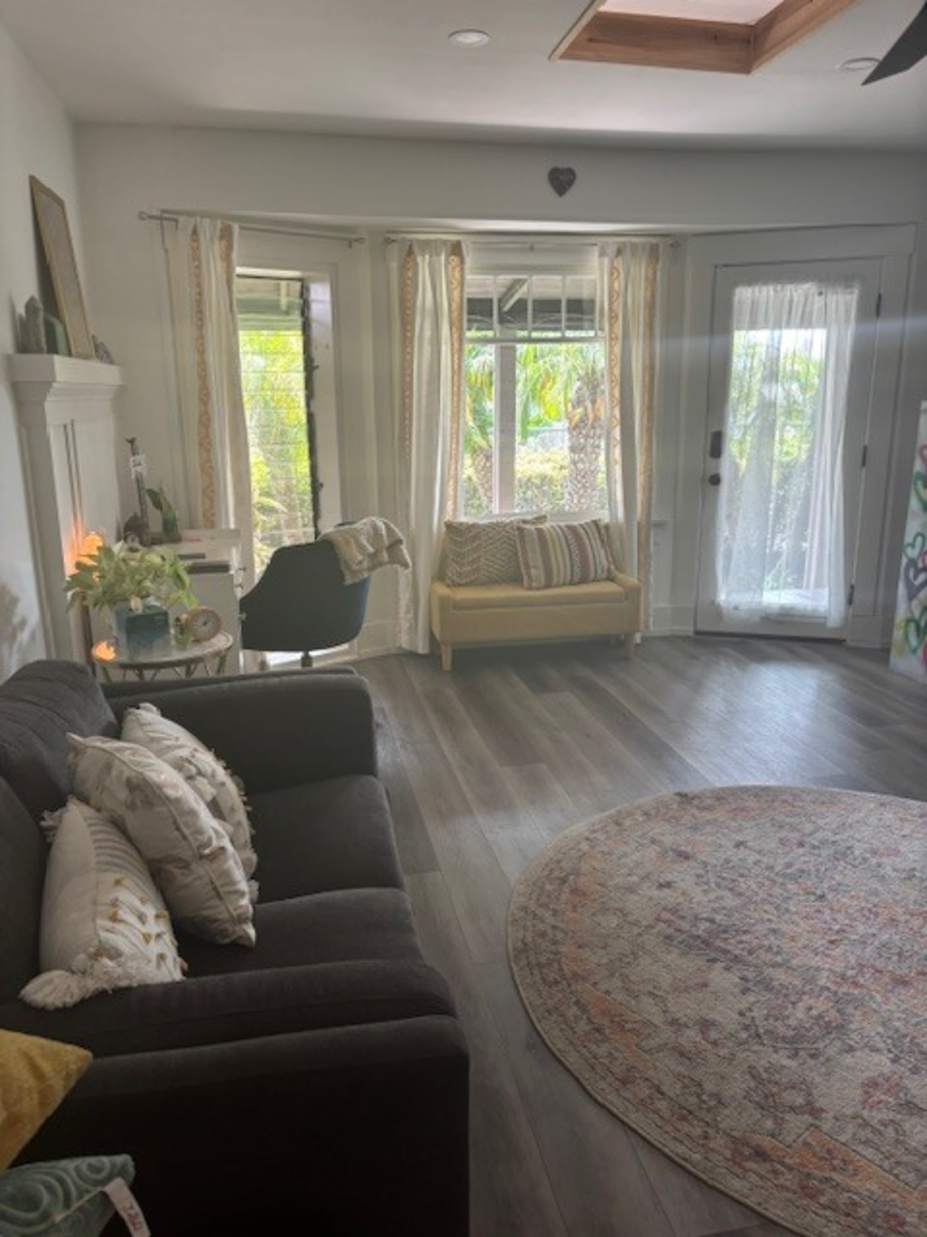 The image shows a spacious living room with a gray sofa, a yellow loveseat, a large round rug, and windows that provide natural light and a view of greenery outside.
