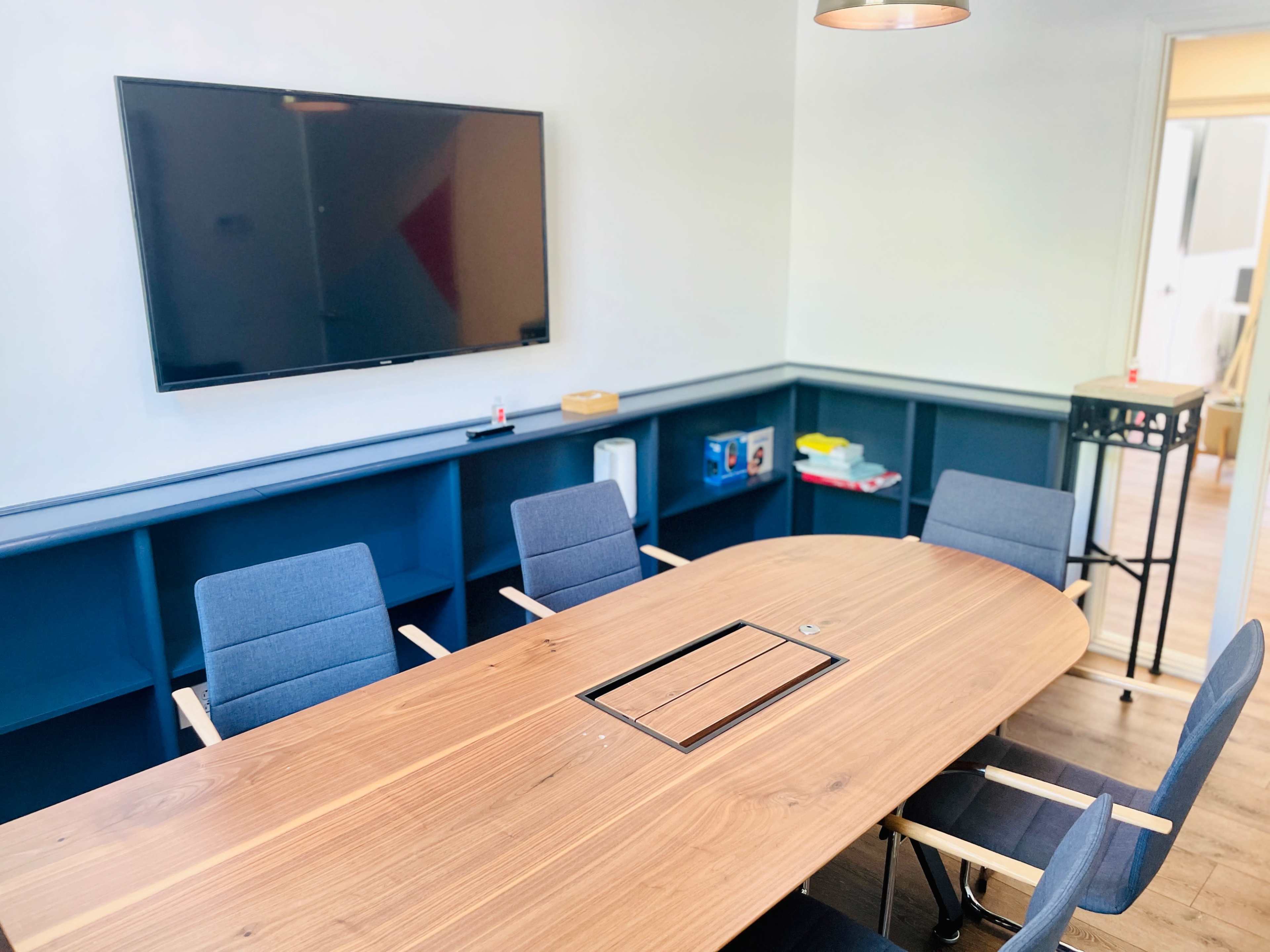 A modern conference room features a large wooden table surrounded by gray chairs and a wall-mounted television.