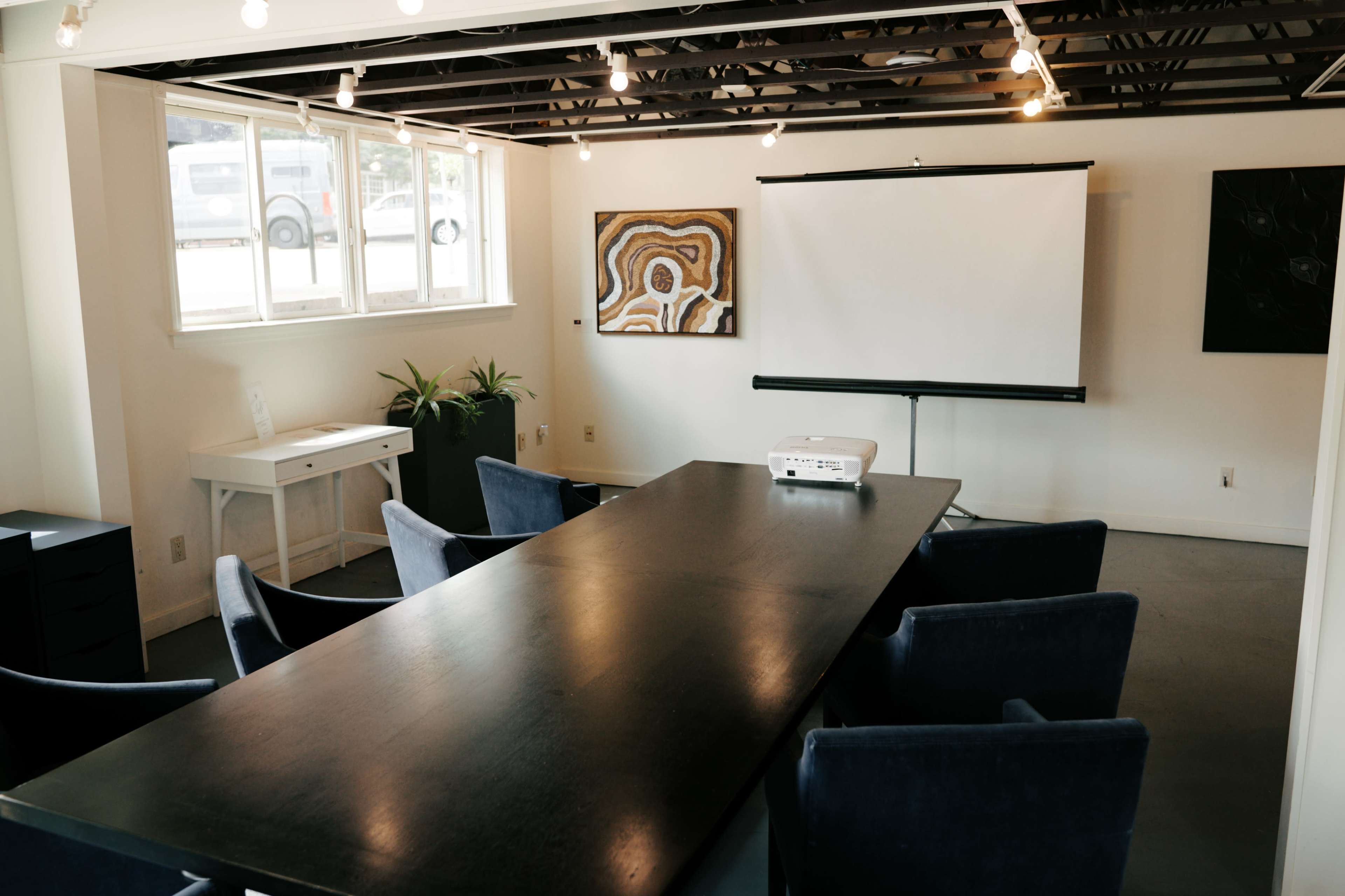 The image shows a conference room featuring a large black table, blue chairs, a projector on the table, and a blank projection screen mounted on a stand against the wall.