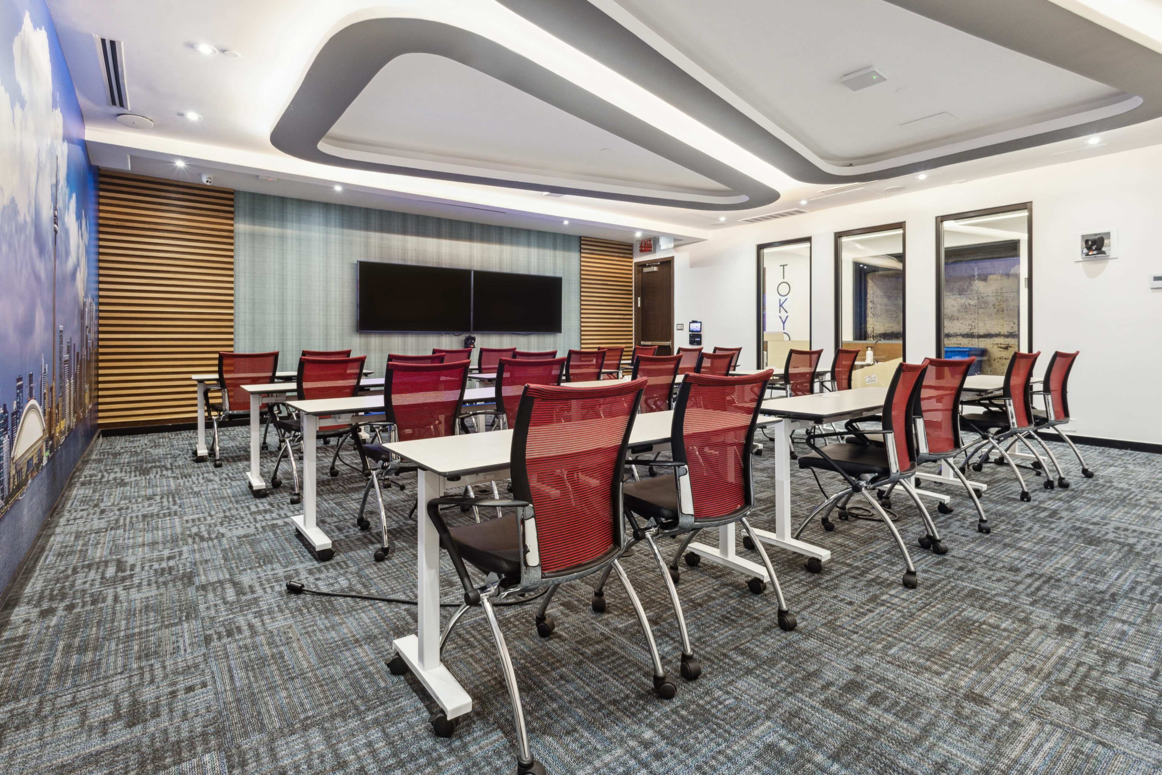 The image showcases a contemporary meeting room featuring rows of red and white chairs arranged around tables, with a large screen on the wall and windows reflecting the cityscape.