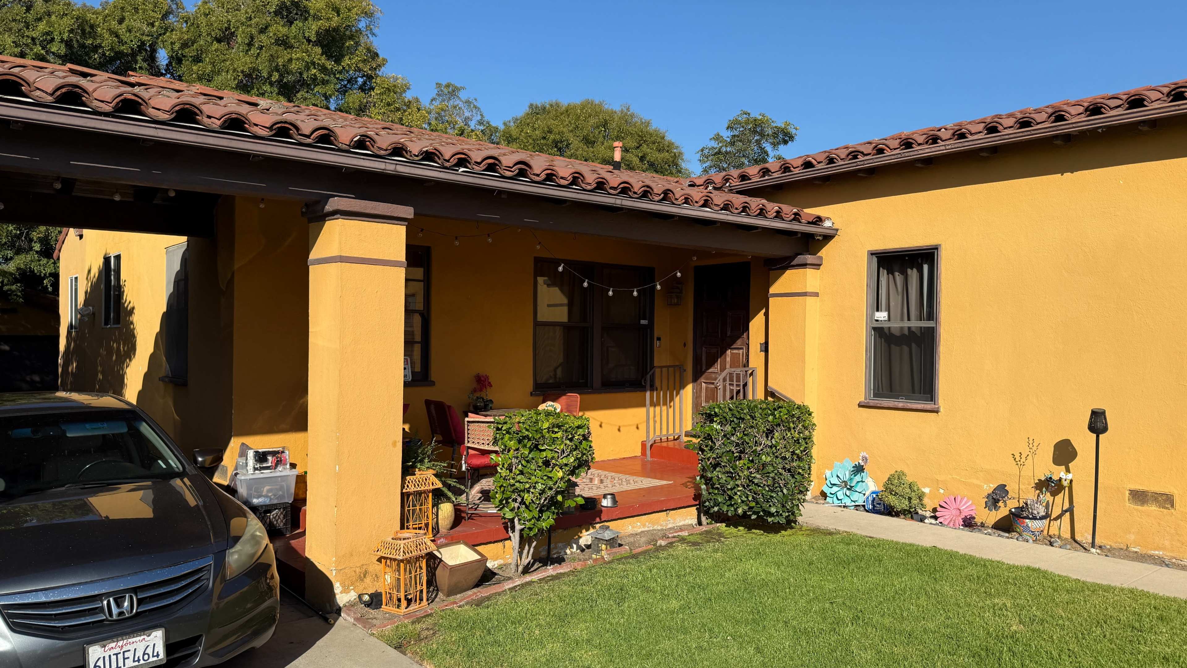 The image shows a yellow house with a tiled roof, featuring a front porch with seating and decorative elements, alongside a parked gray car in the driveway.