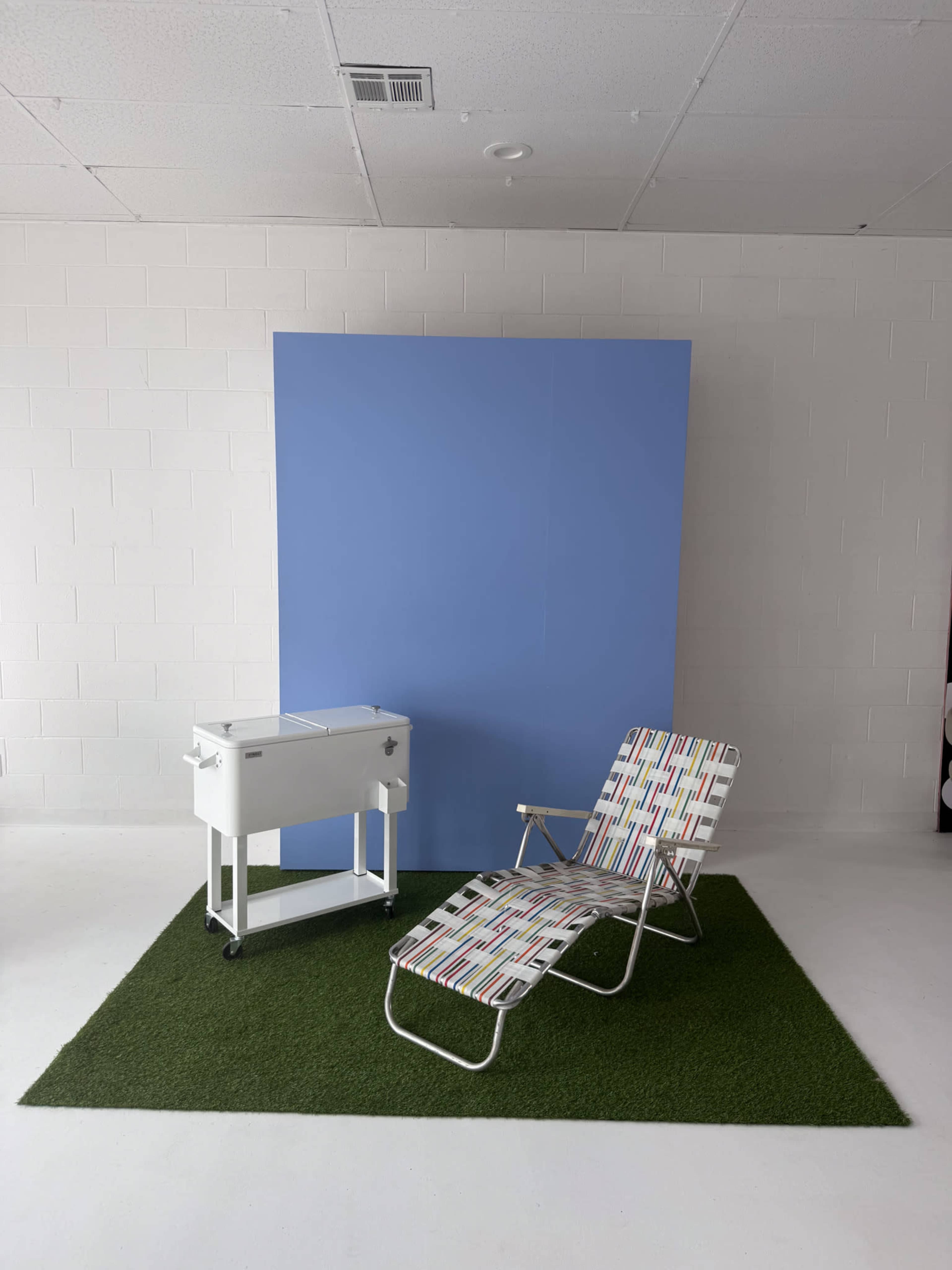 A white rolling cart and a folding lounge chair are positioned on a green rug in front of a blue backdrop in a minimalistic indoor setting.