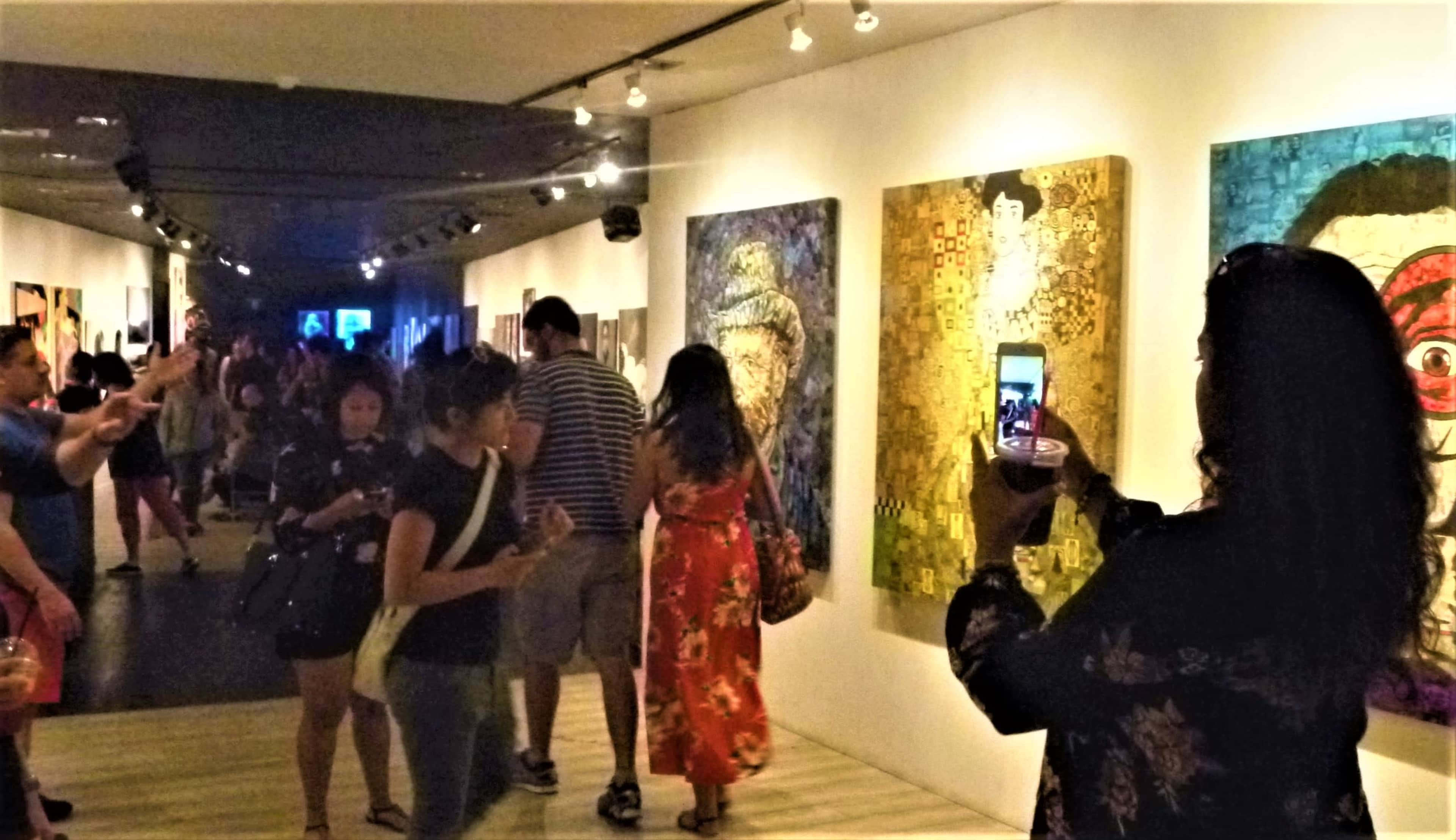 A group of visitors explores an art gallery, with some individuals viewing paintings while others take photos.