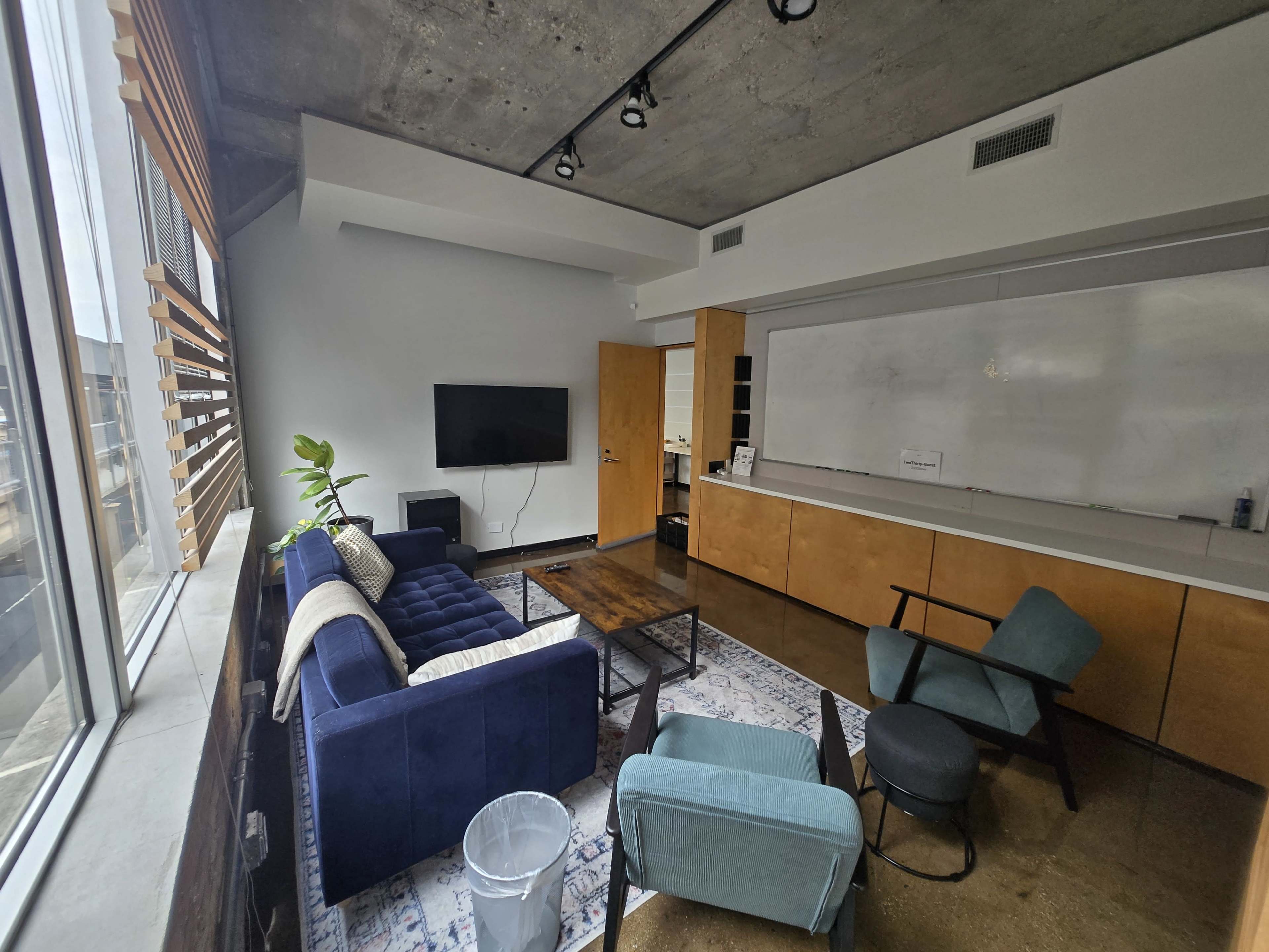 The image shows a modern office space with a blue sofa, a coffee table, and a television mounted on the wall, illuminated by natural light filtering through large windows.
