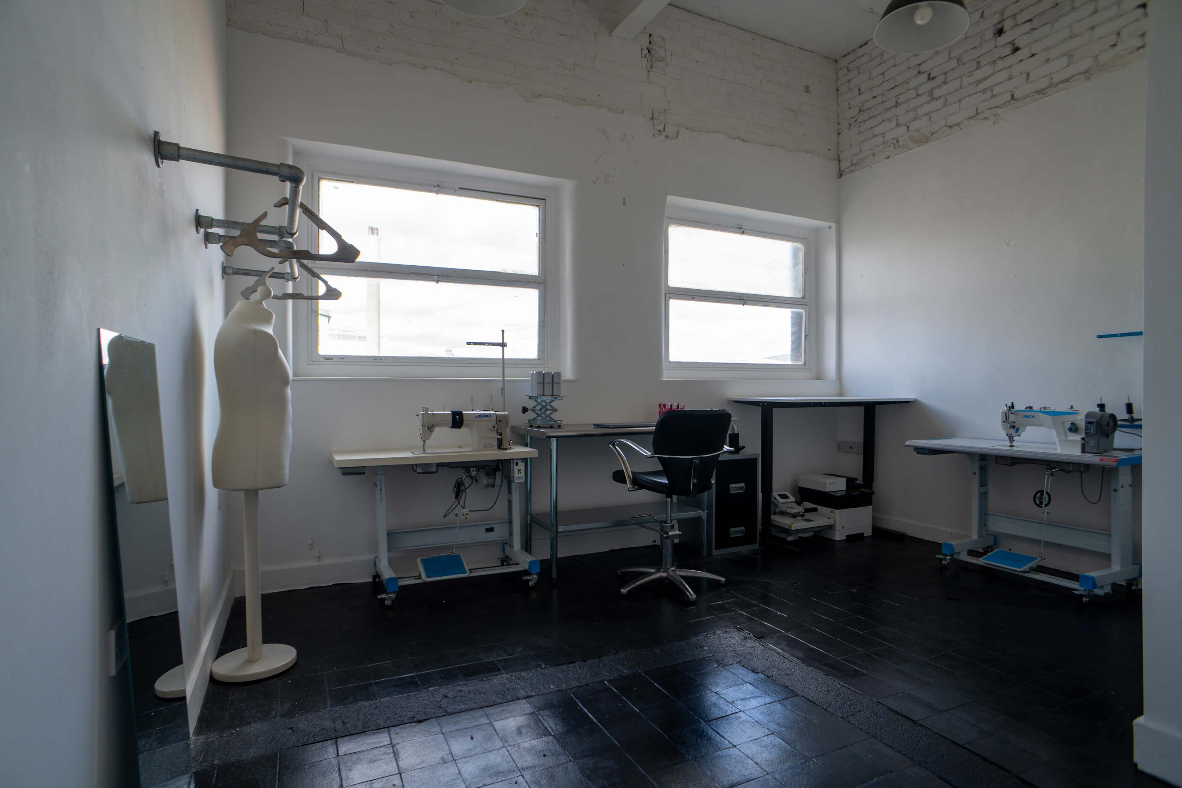 The image shows a bright room with two sewing tables, a chair, a dress form, and a mirror against a white brick wall.