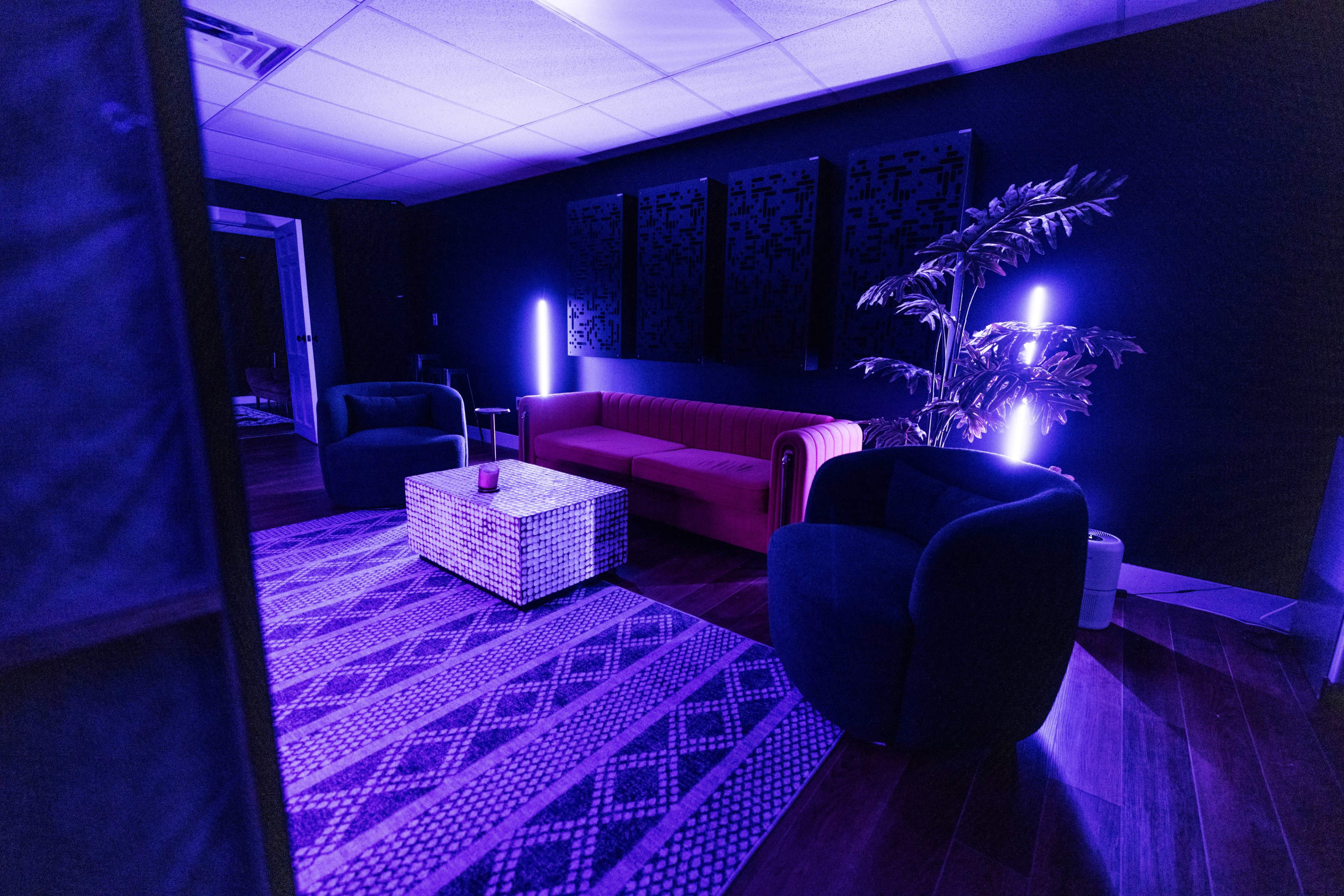 A modern lounge area features a red sofa, two blue armchairs, and a patterned rug under purple lighting.