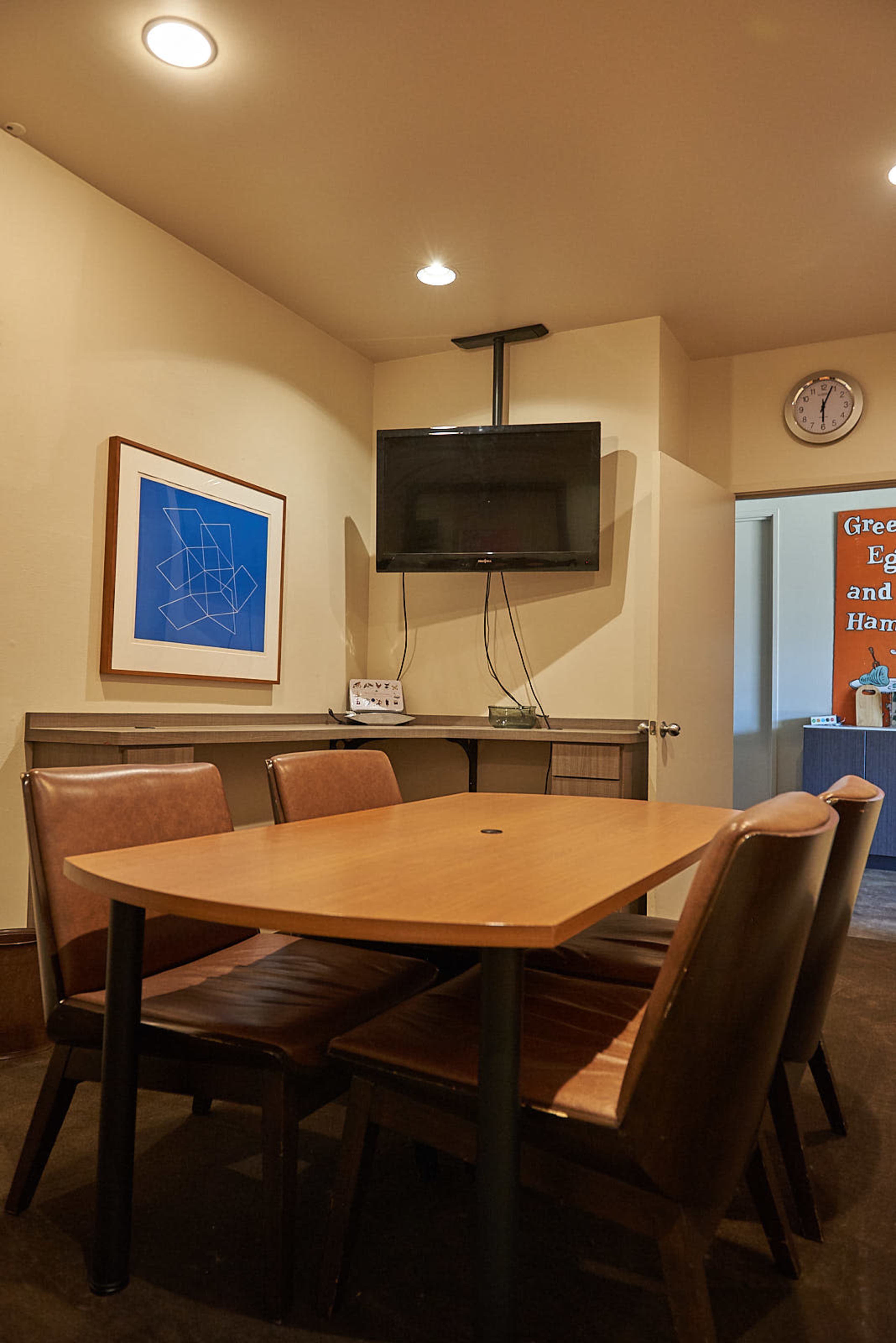 A small meeting room features a table with four chairs, a wall-mounted television, and a clock on the wall.