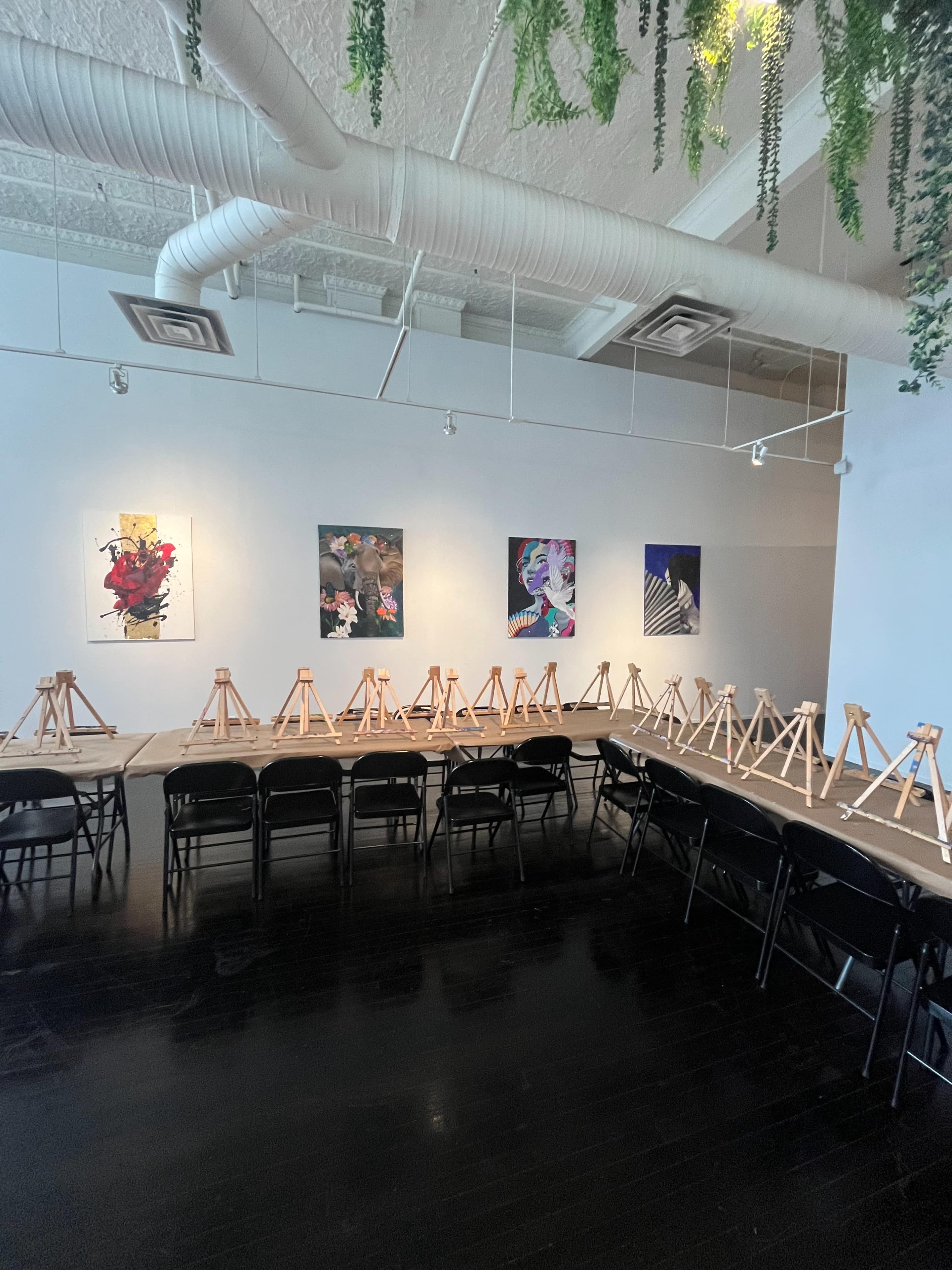 The image shows an art studio with several easels arranged in rows facing a wall displaying colorful artwork.