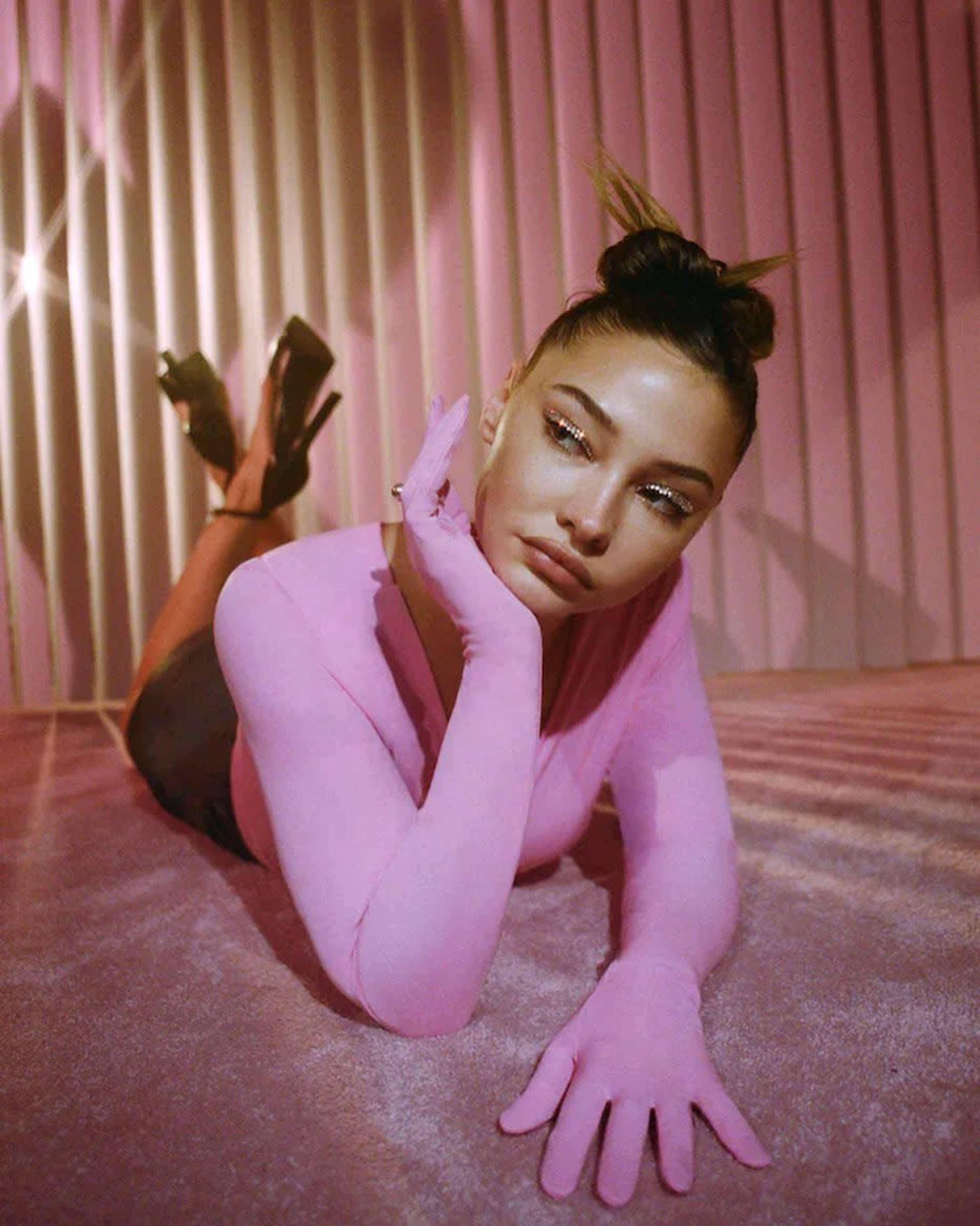 A person lies on a pink surface, wearing a long-sleeved pink top and high heels, with a hand resting on their chin against a backdrop of vertical stripes.
