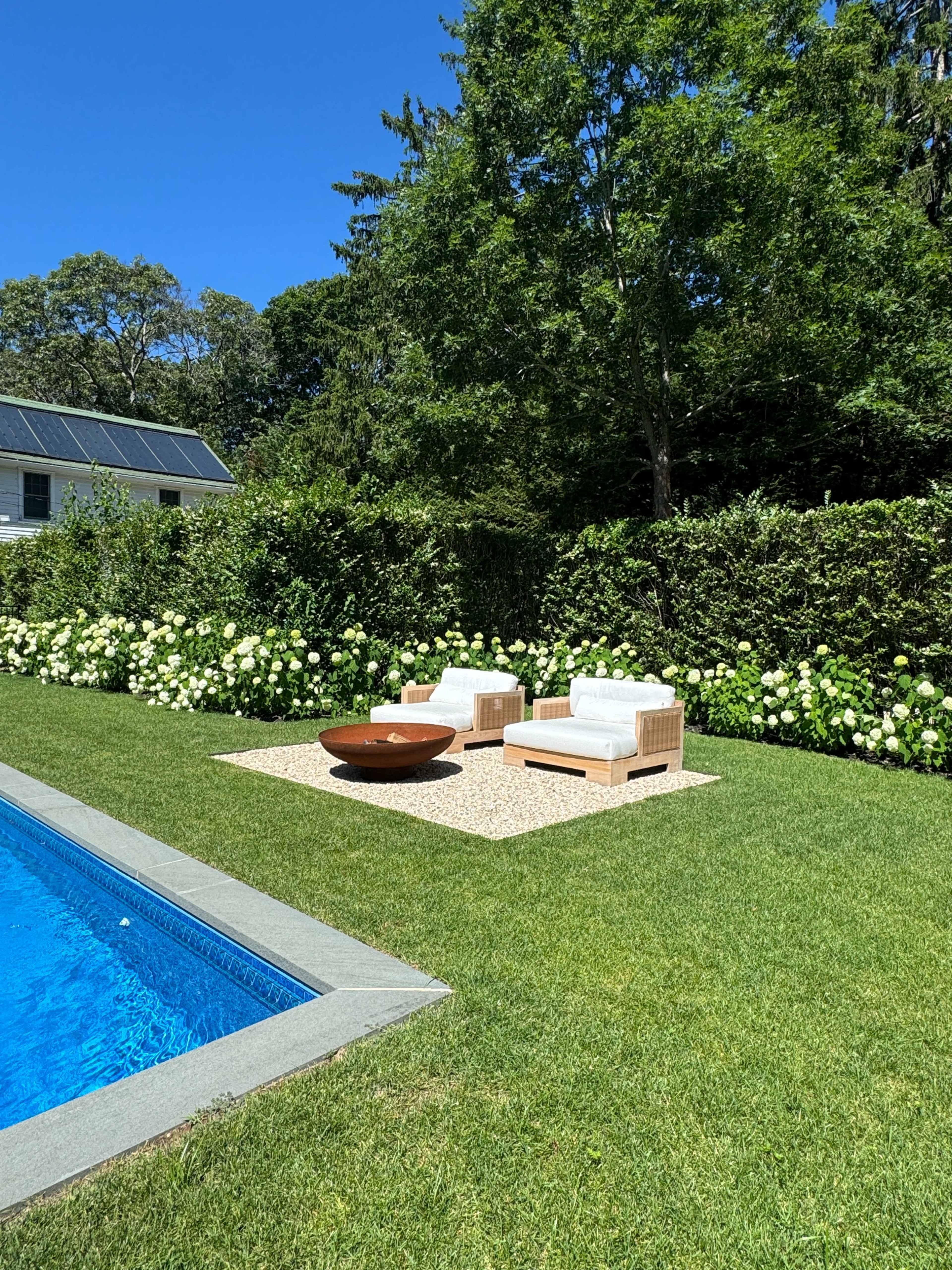 A poolside area with two white chairs and a round table surrounded by green grass and flowering bushes.