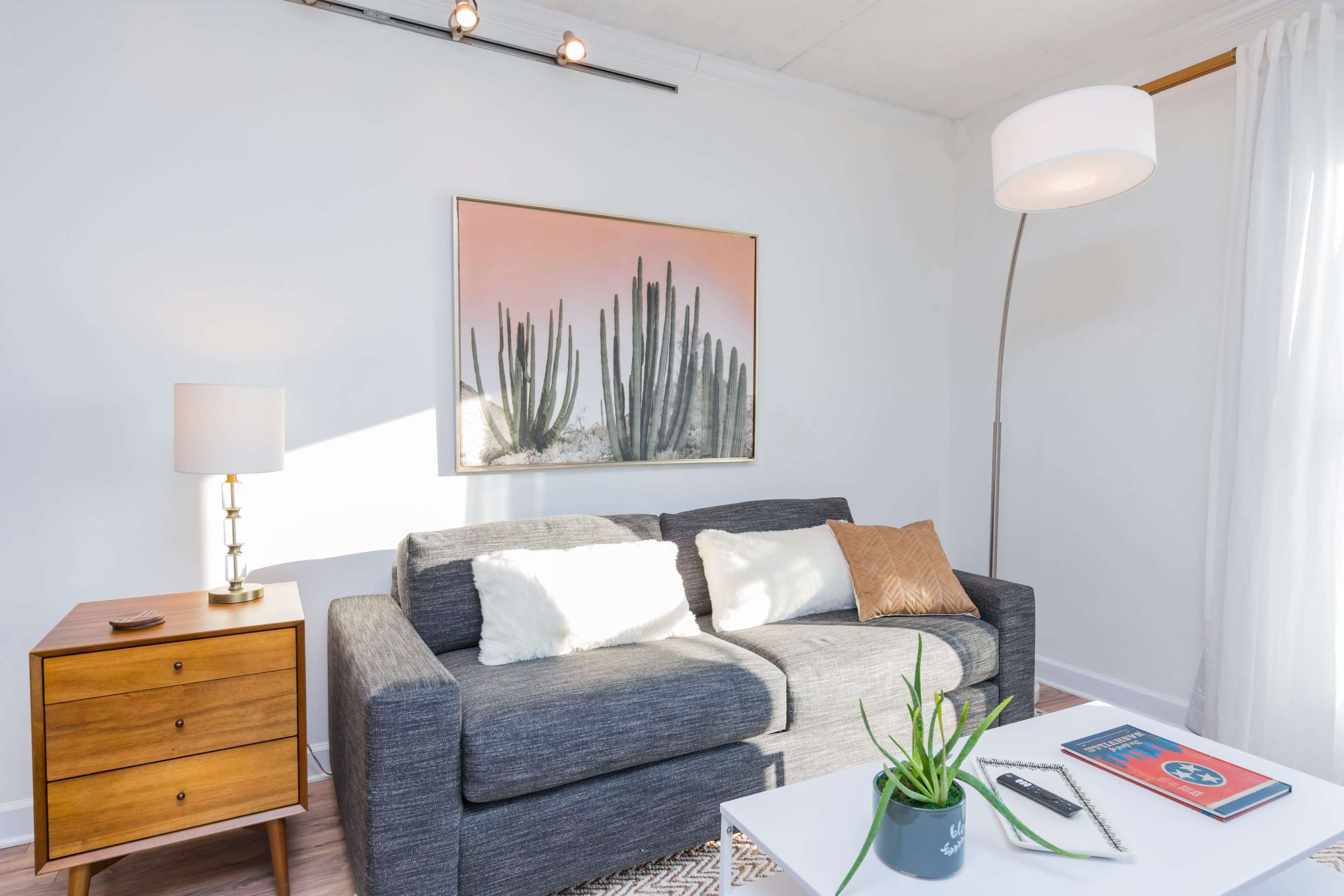 The scene features a modern living room with a gray sofa, a wooden side table with a lamp, and a wall art piece depicting cacti.