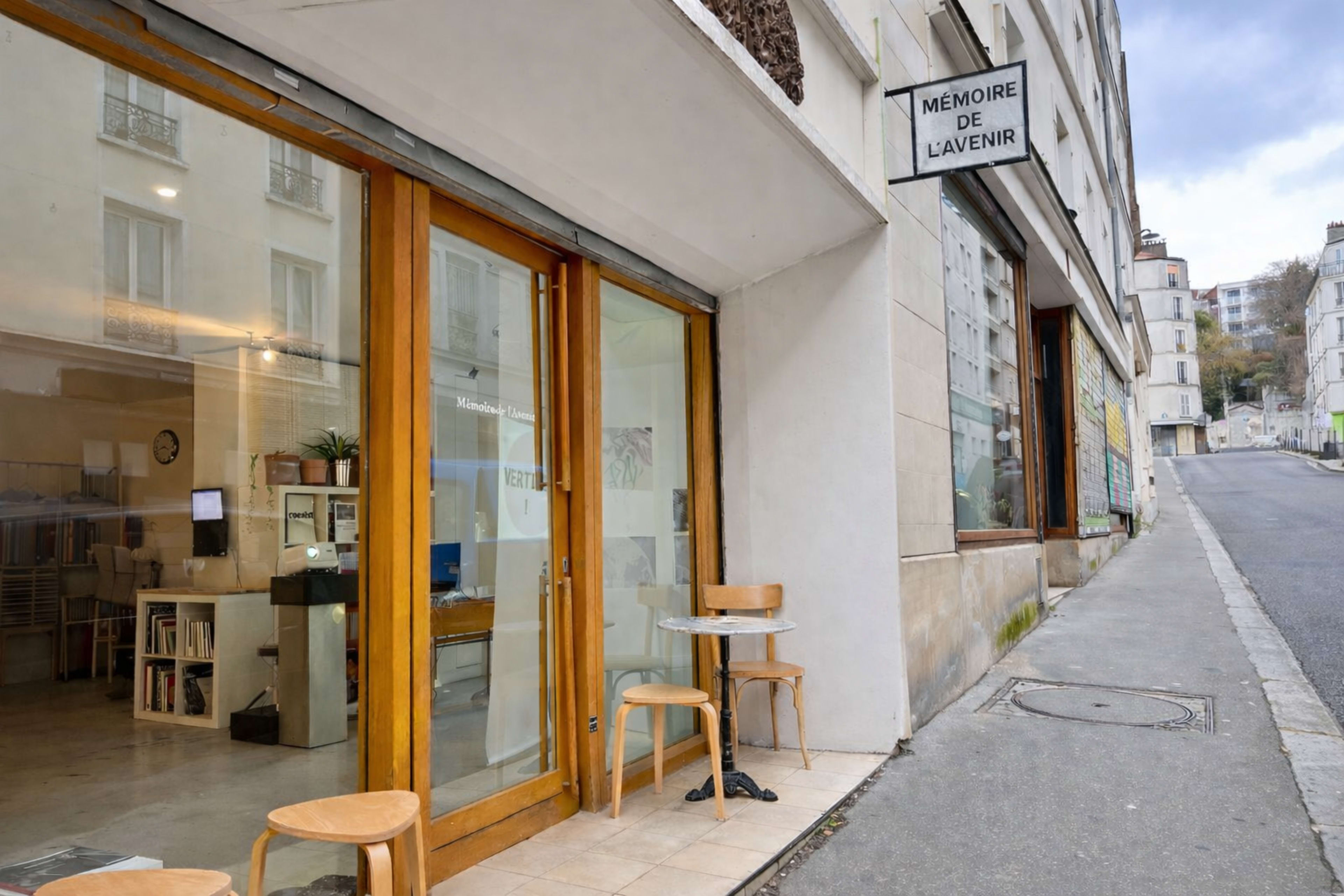 The image shows a storefront with large glass doors and wooden chairs outside, situated on a sloped street.