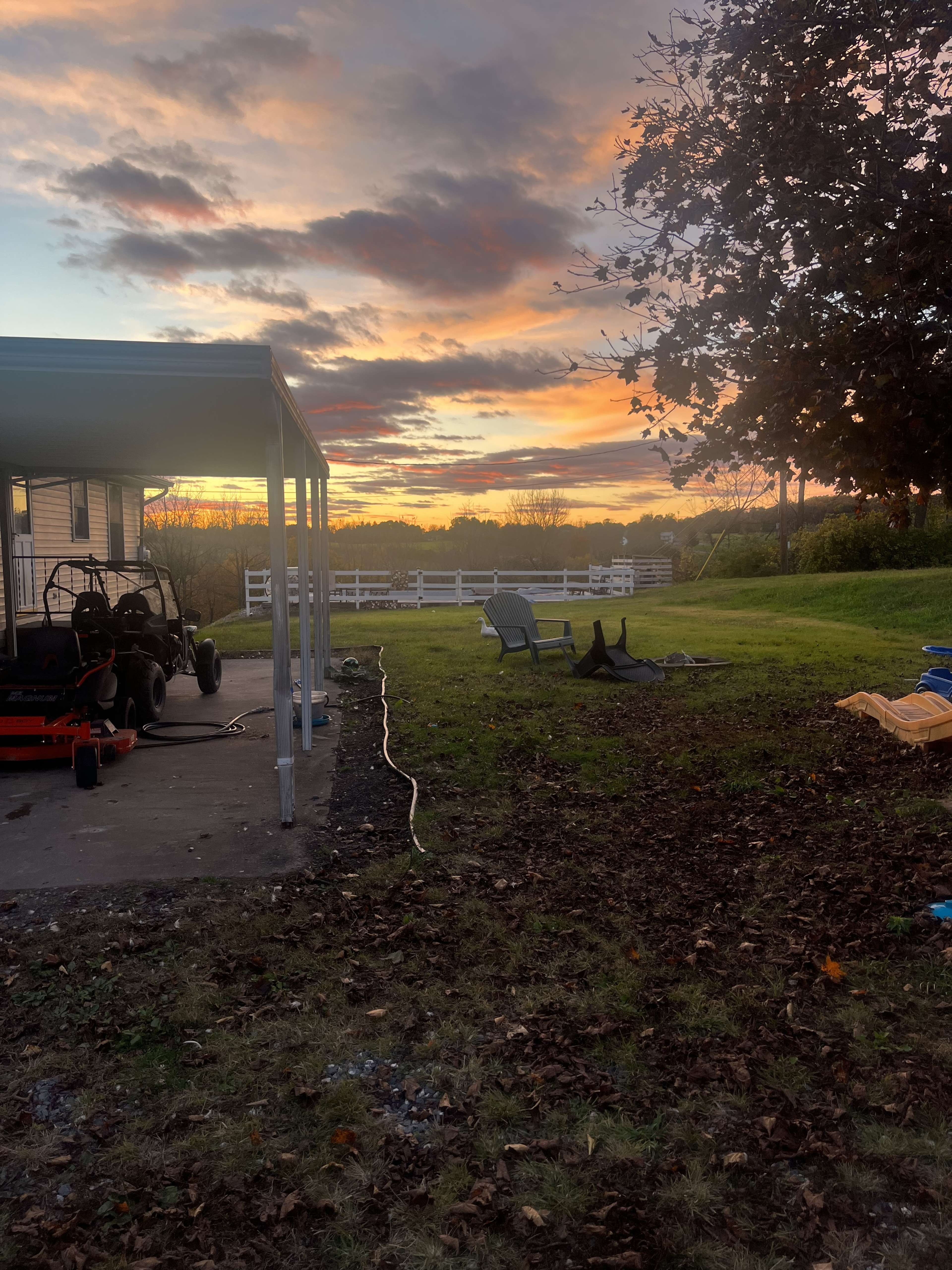 The scene shows a backyard with a parked lawnmower under a covered area, chairs positioned on the grass, and a colorful sunset in the background.