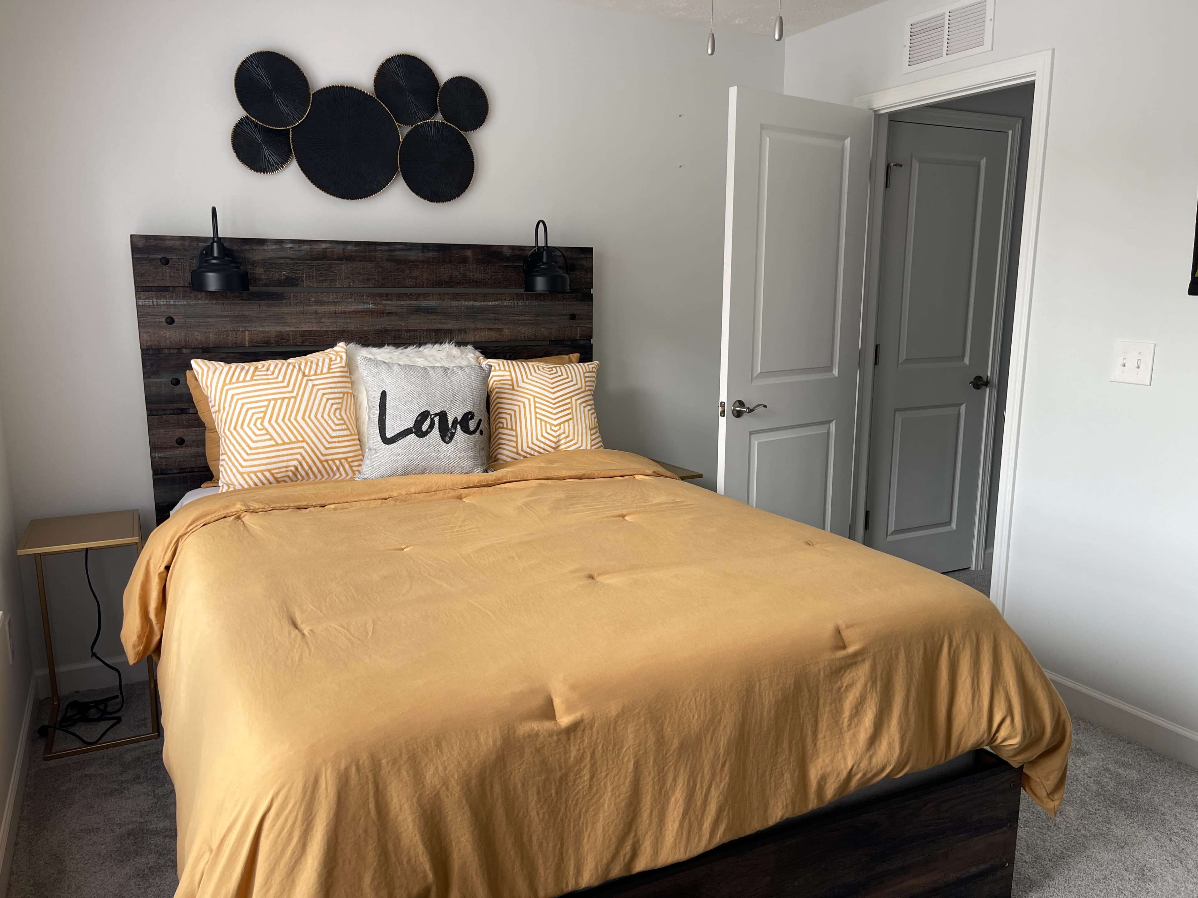 A well-lit bedroom featuring a wooden bed frame with a yellow comforter, two patterned pillows, and a decorative cushion that reads "Love," along with a nightstand and a closed door.