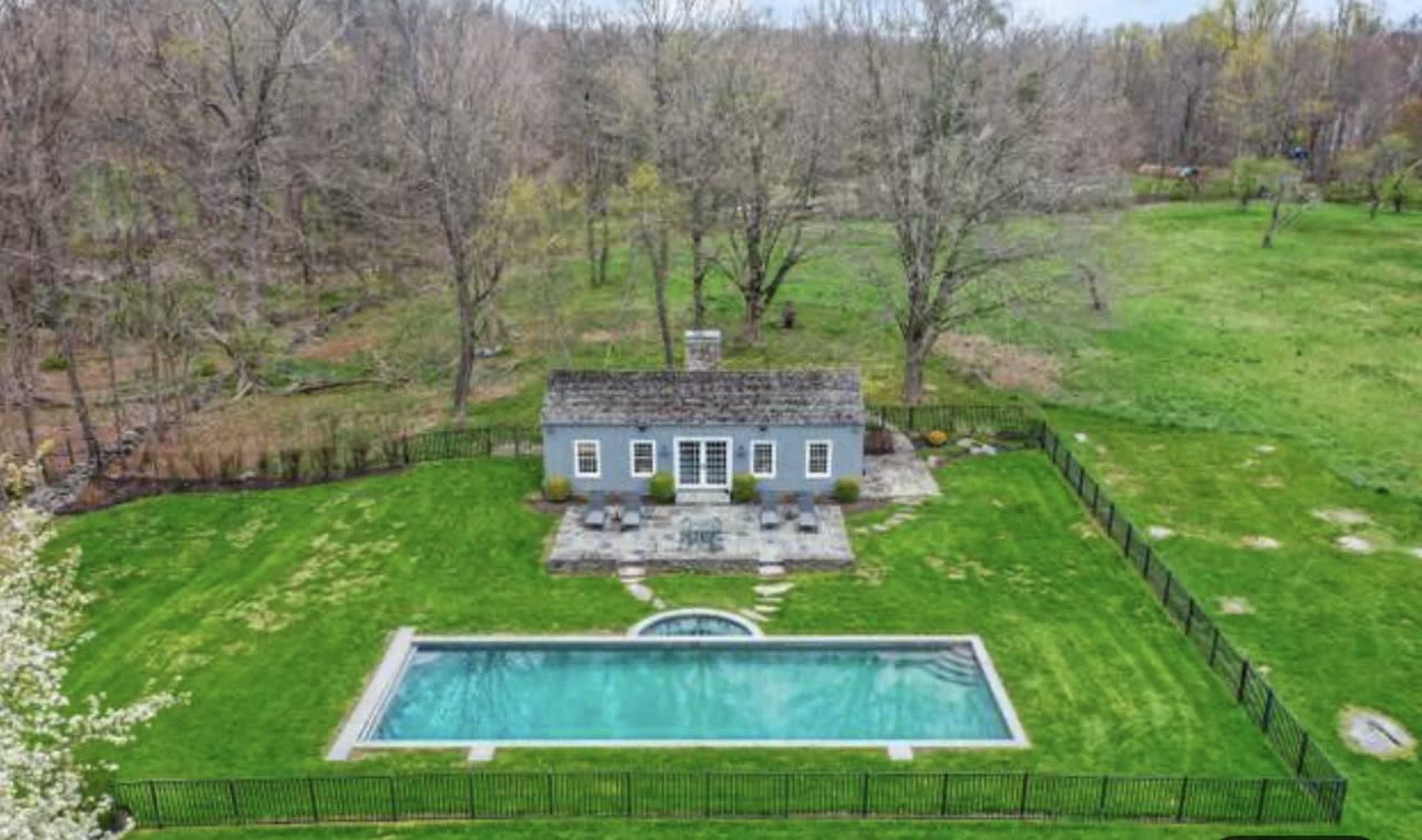 A backyard featuring a swimming pool, a small house, and a large green lawn surrounded by trees.
