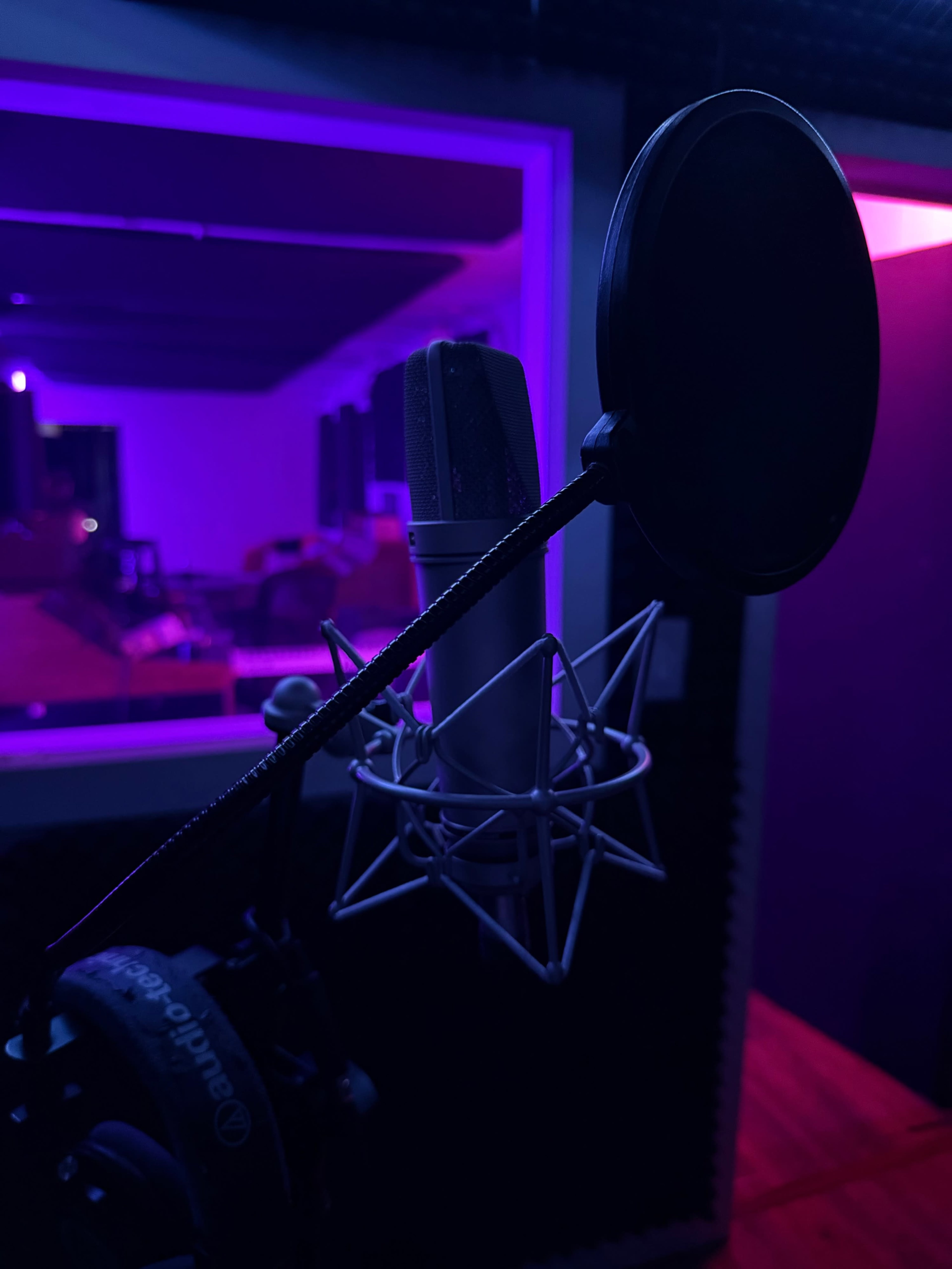 A microphone with a pop filter is positioned in a dimly lit recording studio, illuminated by purple and pink lights.