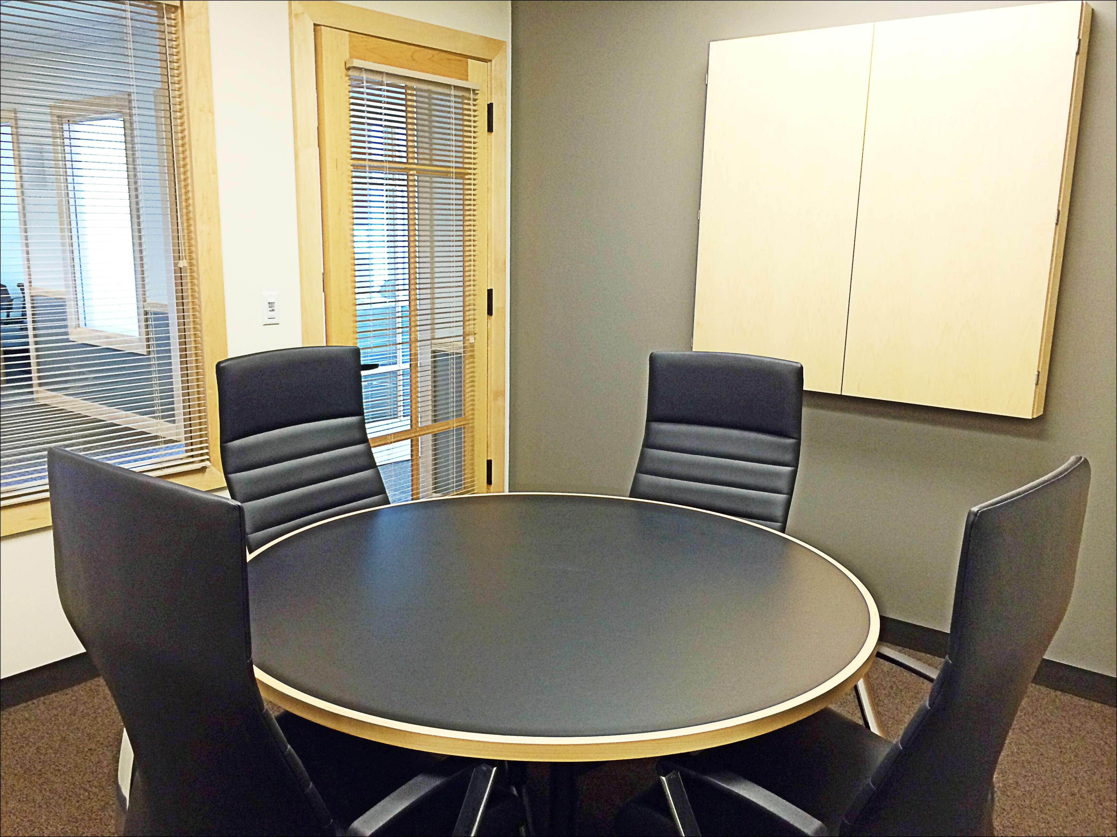 A circular conference table is set in a small meeting room with four black chairs and a wall-mounted cabinet.