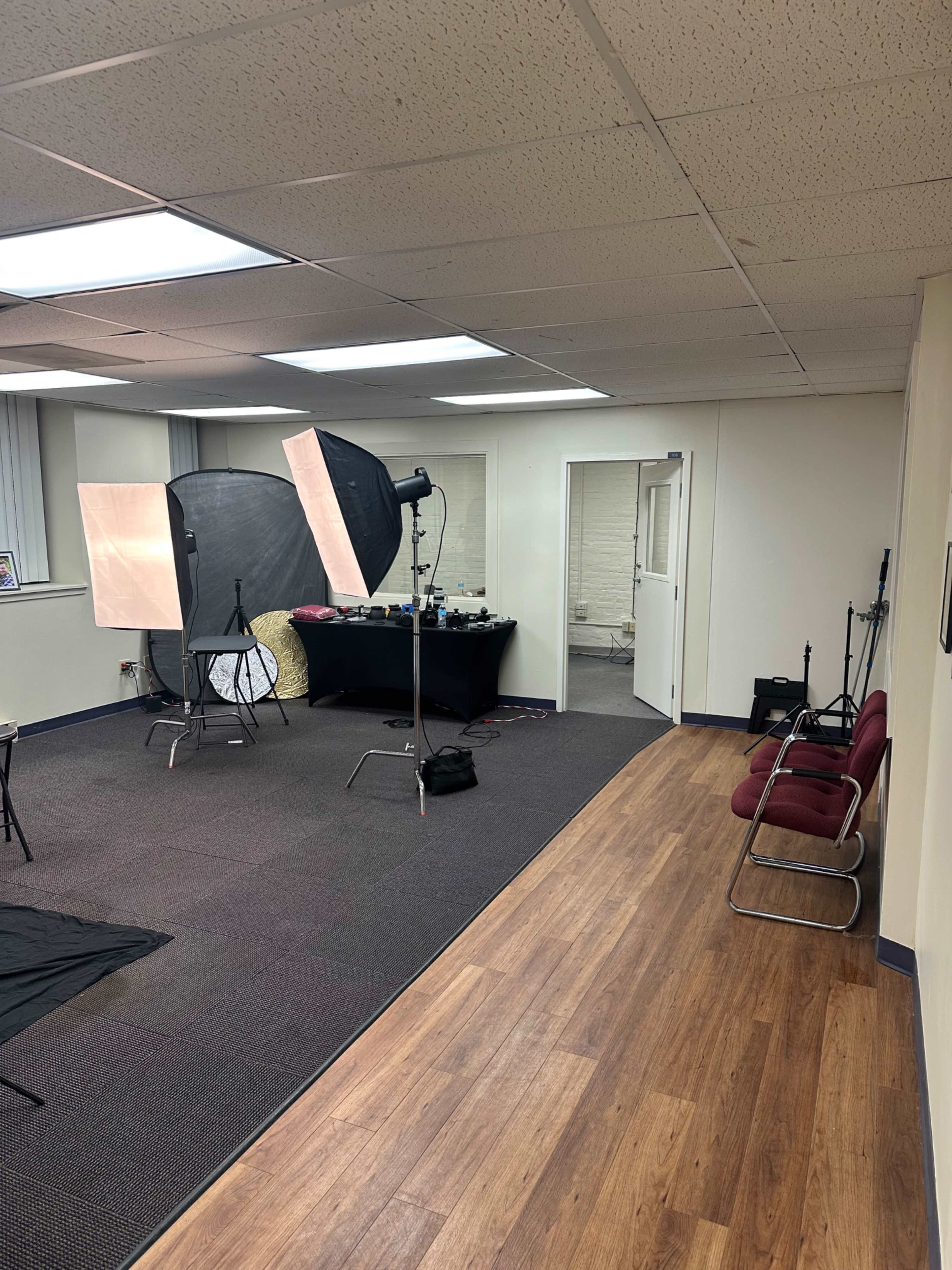 The image shows a photography studio setup with lighting equipment and a table in one corner, while the opposite side features a pair of red chairs.
