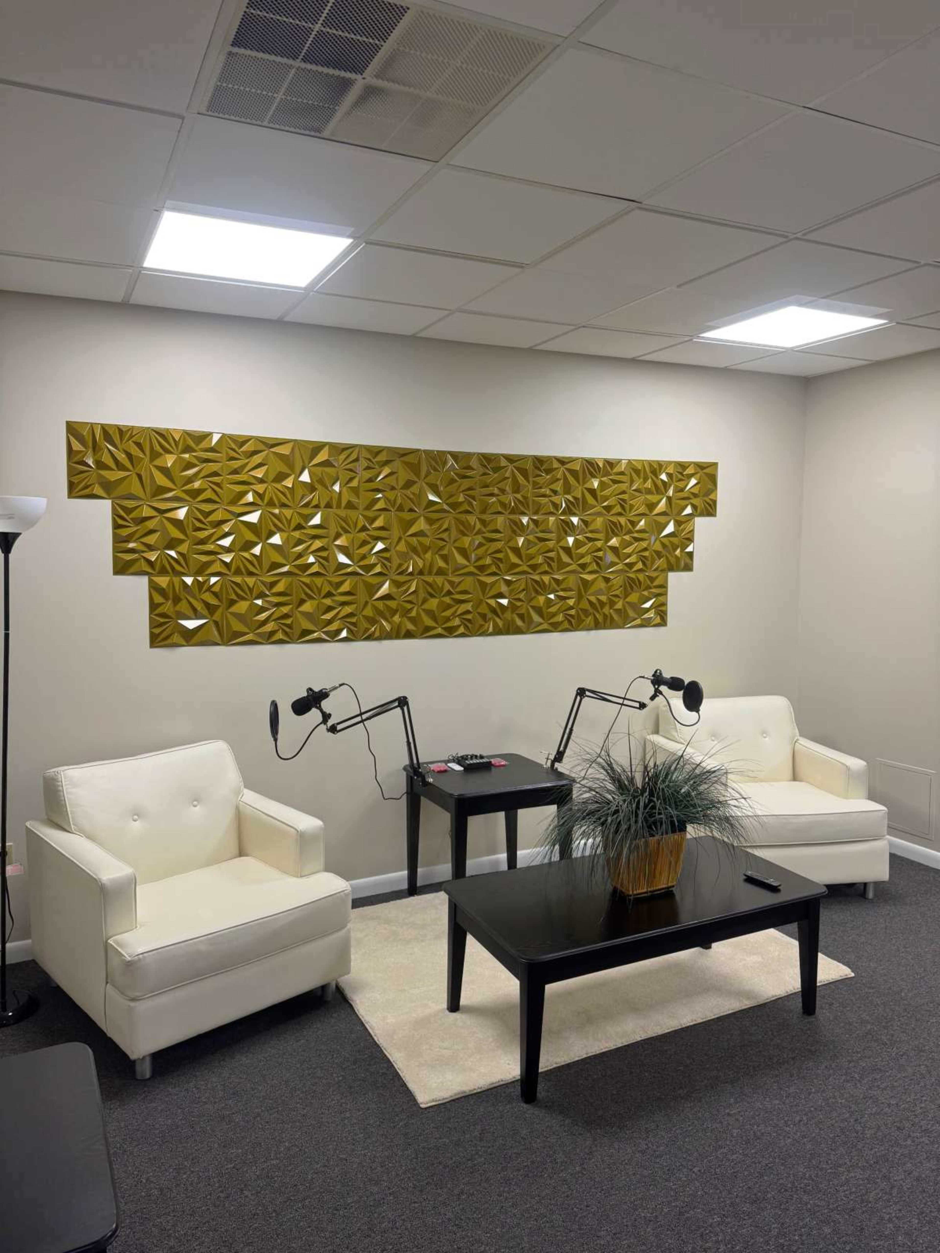 The image shows a minimalist office lounge area with two white armchairs facing a black coffee table, under bright ceiling lights, and featuring a decorative yellow wall panel.