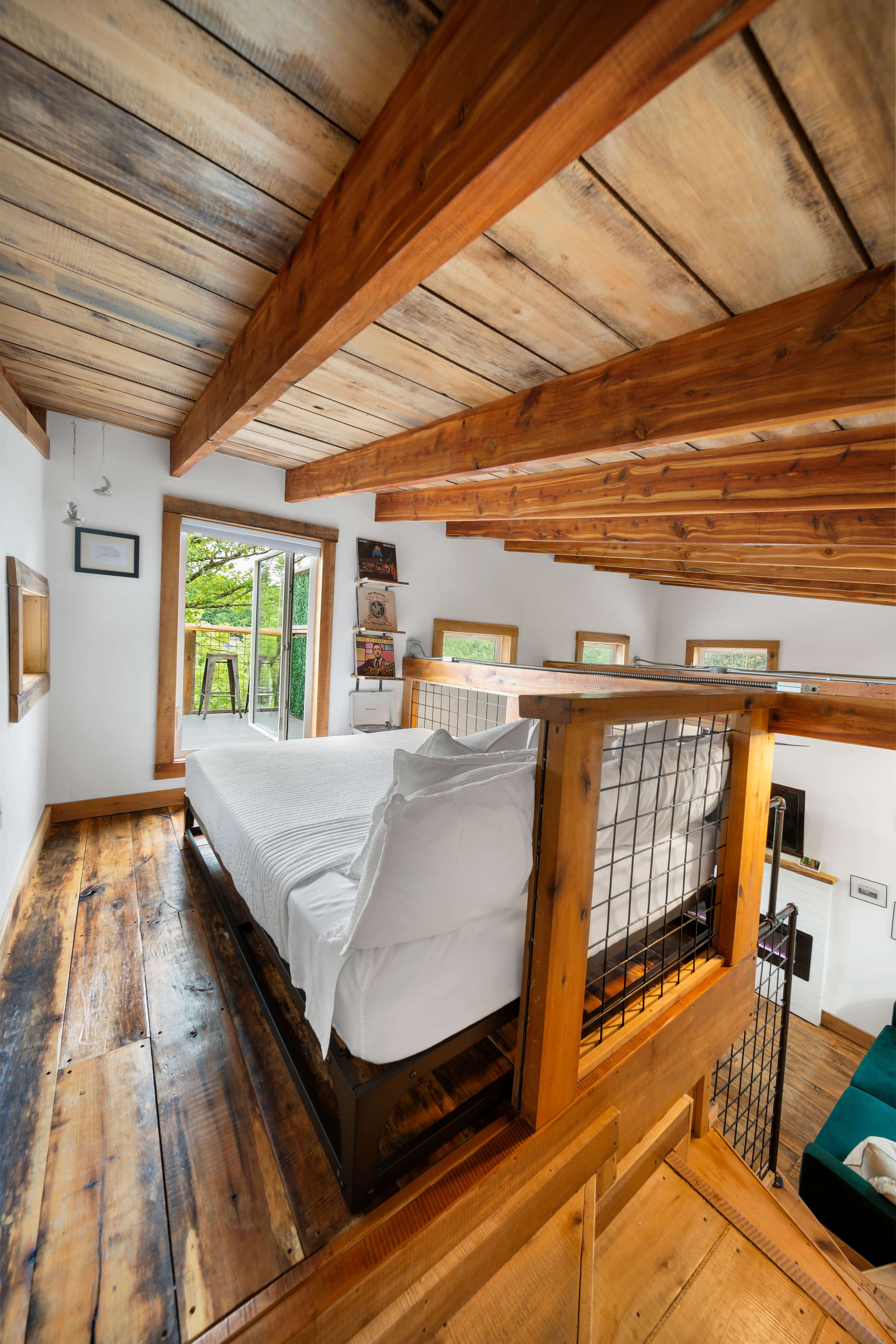 A loft-style bedroom with exposed wooden beams, a white bed, and large windows overlooking greenery.